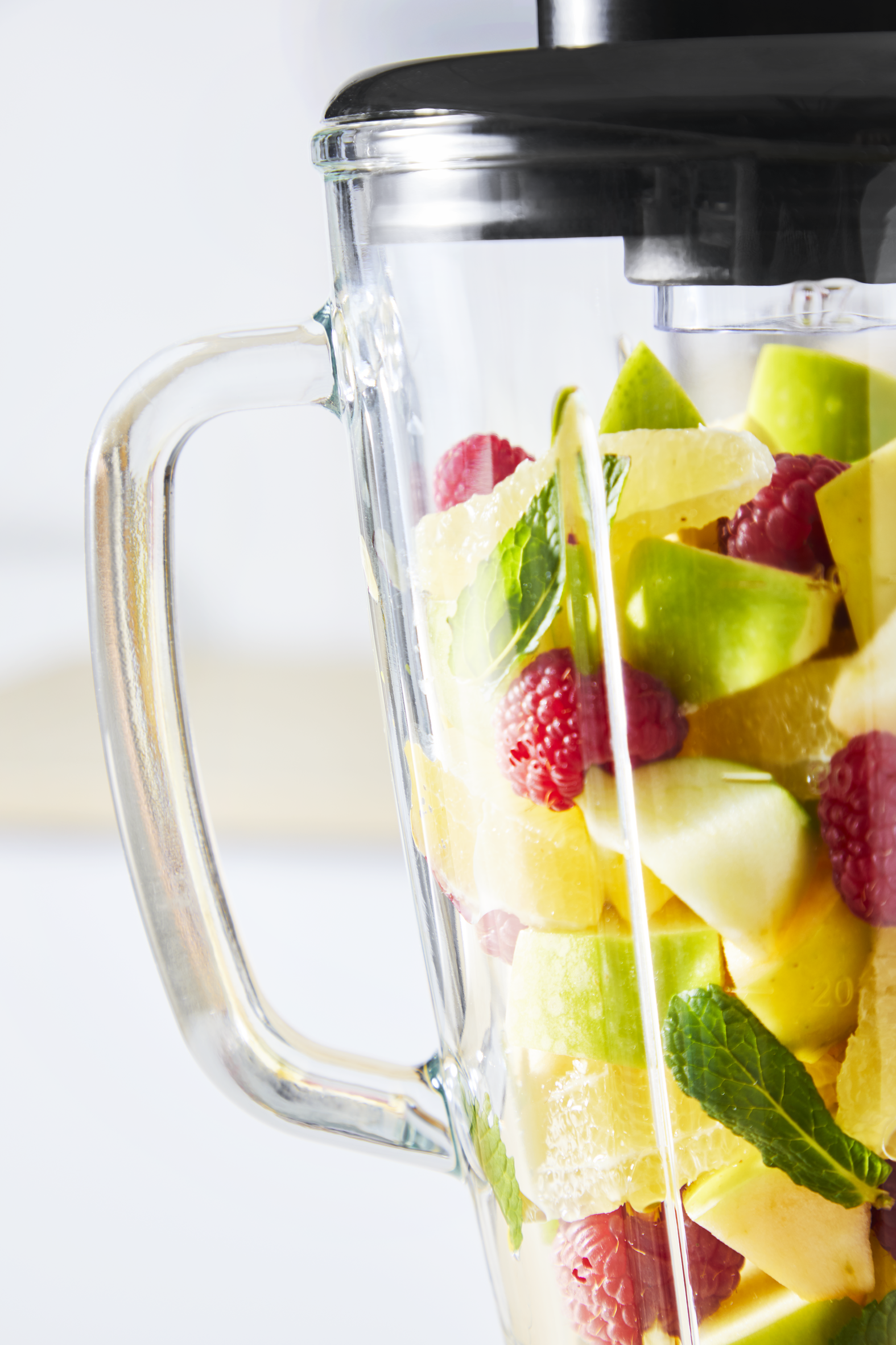 Blender filled with chopped fruit: green apples, raspberries, and citrus fruits. Glass handle visible. White background.