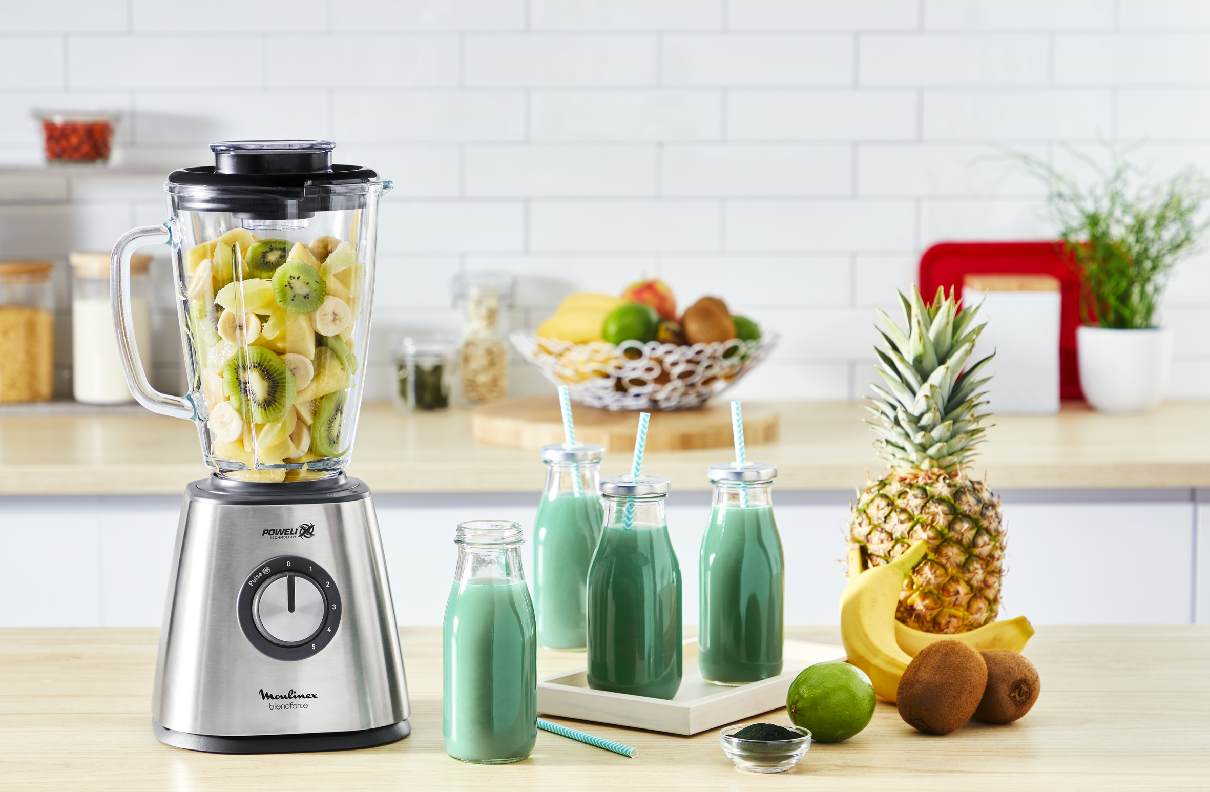 Blender with fruit, juice bottles, and pineapple on a table.