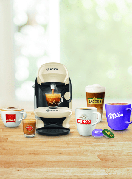 Kaffeemaschine mit Kaffeetassen, darunter Jacobs, Kenco, Milka und kleinere Tassen. Holzoberfläche, unscharfer Hintergrund.