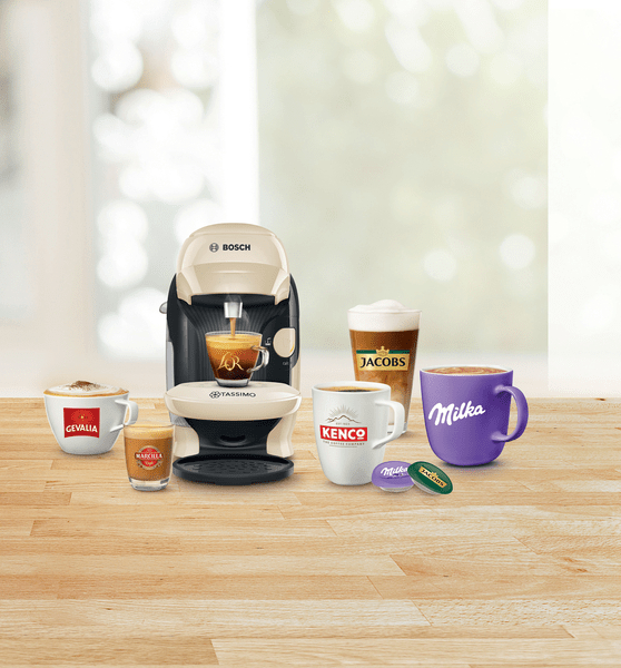 Eine Kaffeemaschine, die Kaffee ausgibt, neben verschiedenen Kaffeegetränken und Kaffeepads auf einem Holztisch.