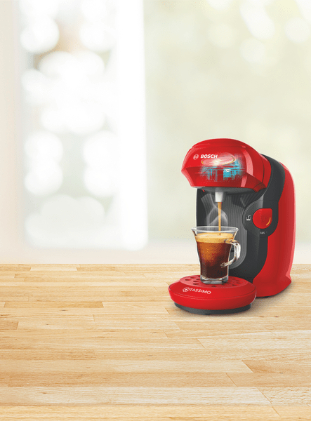 Eine rote Bosch Tassimo Kaffeemaschine, die Kaffee in eine Glastasse auf einer Holzoberfläche ausgibt.