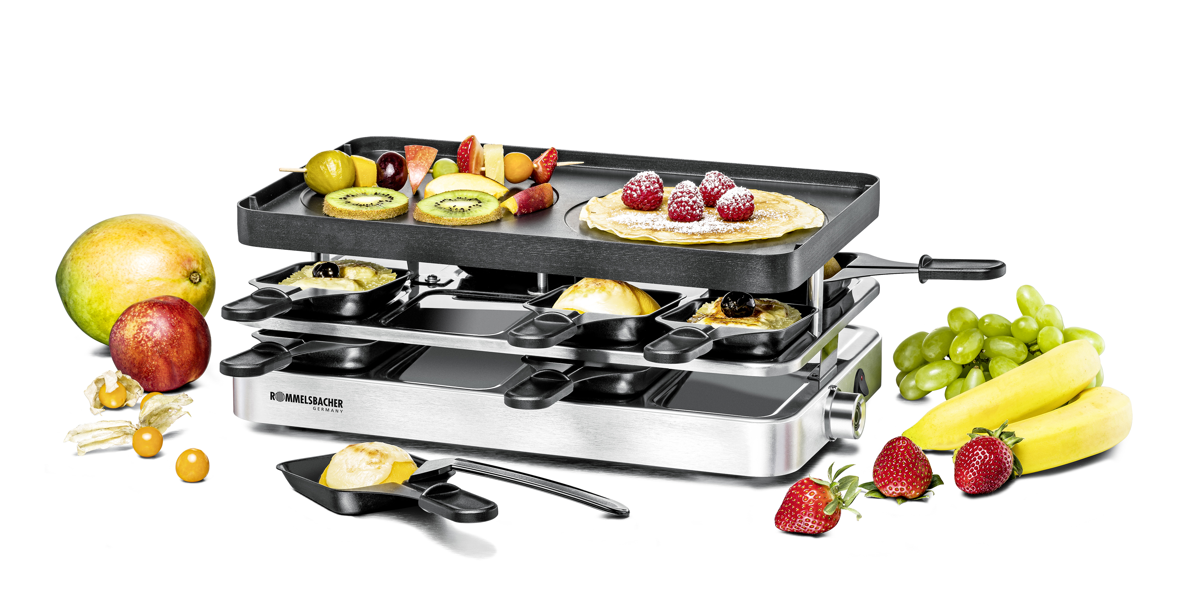 Raclette-Grill mit Essen: Obst, Gemüse und Pfannkuchen. Weißer Hintergrund. Mehrere kleine Pfannen sind drauf.