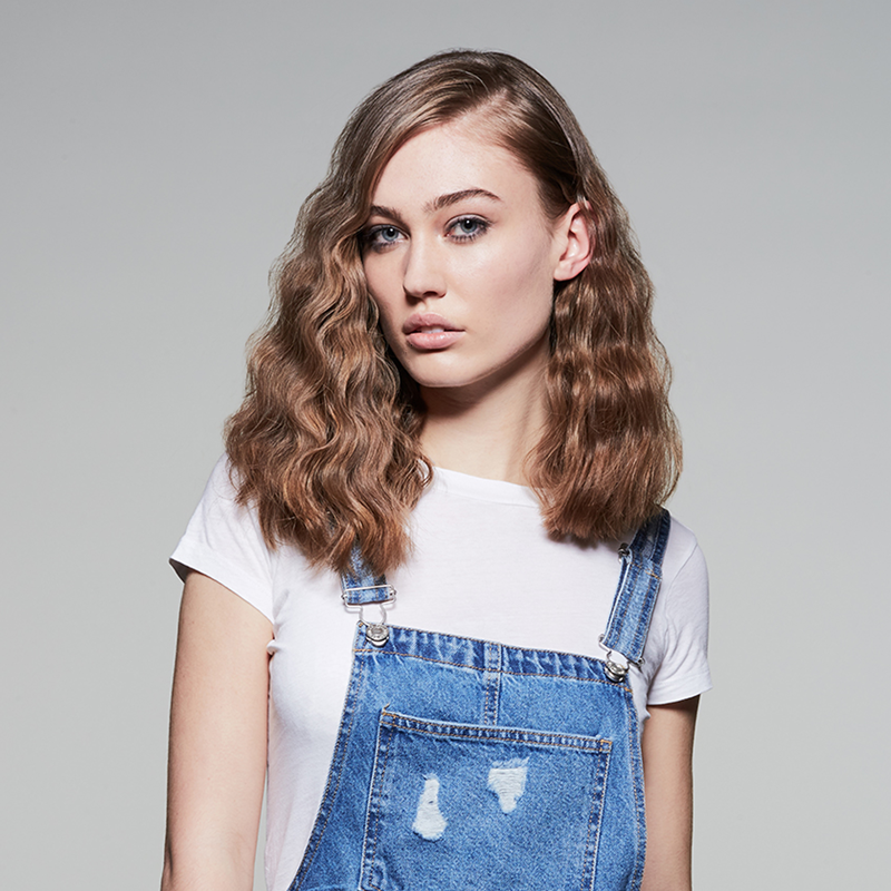 Eine Frau mit welligem Haar in einem weißen Hemd und Jeans-Latzhosen.