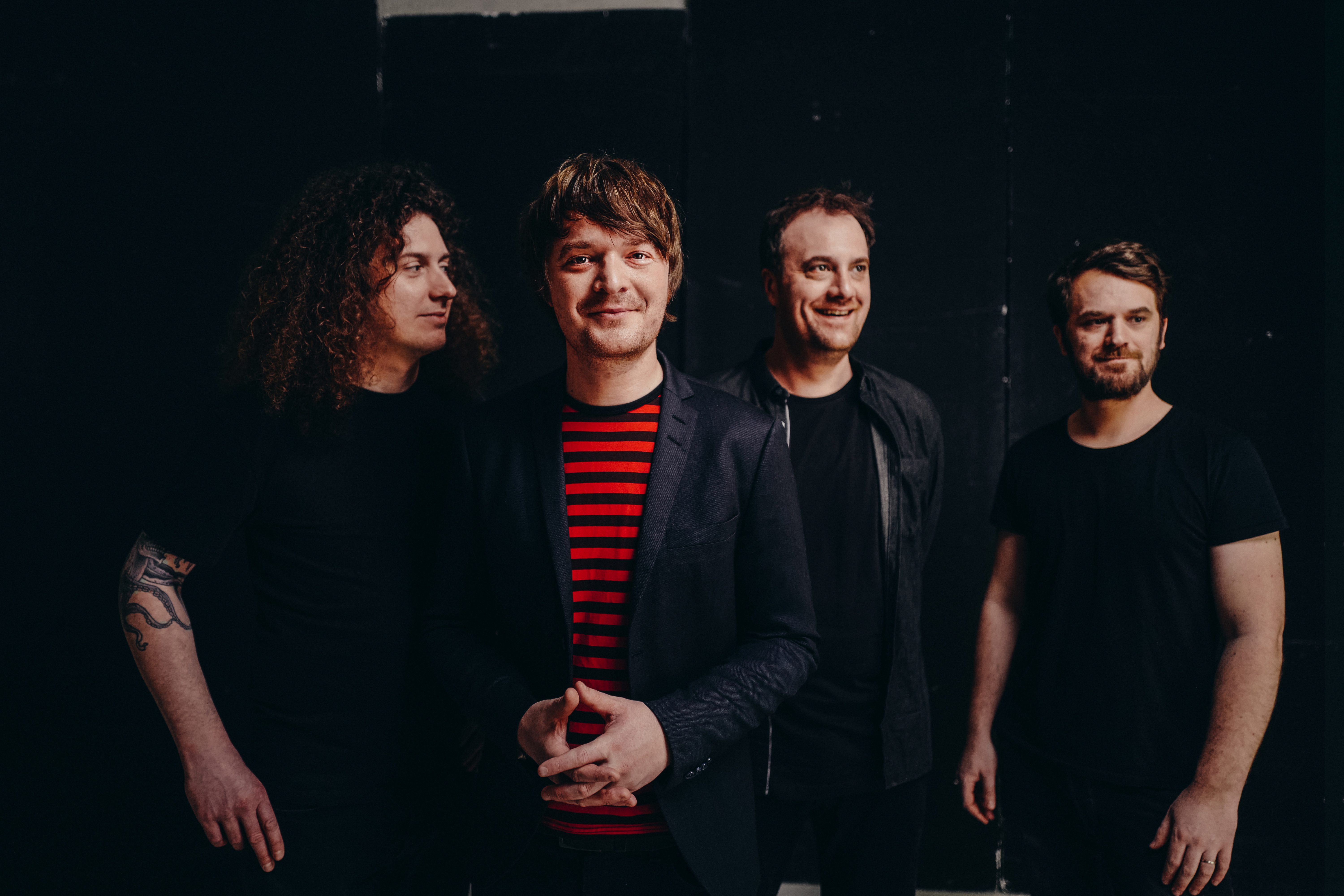 Eine Band aus vier Männern, die vor einem schwarzen Hintergrund posieren. Sie tragen dunkle Kleidung.