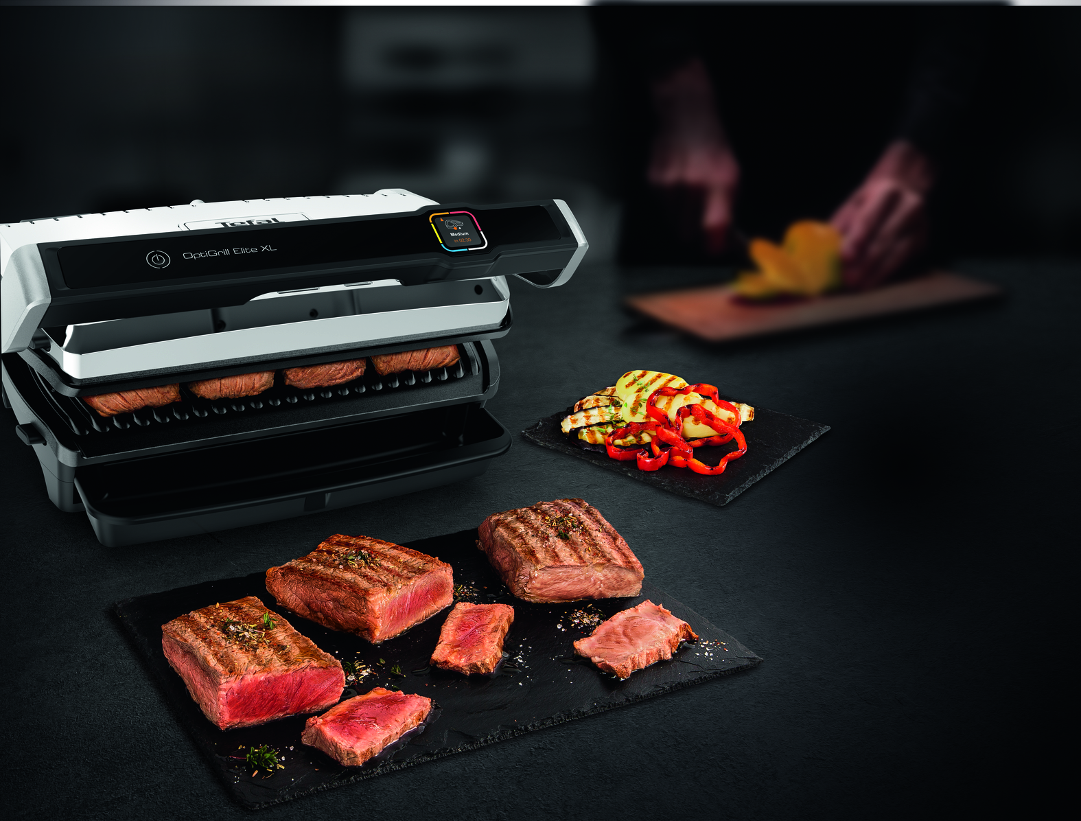 A grill with grilled steaks and vegetables on slate plates, a person in the background.