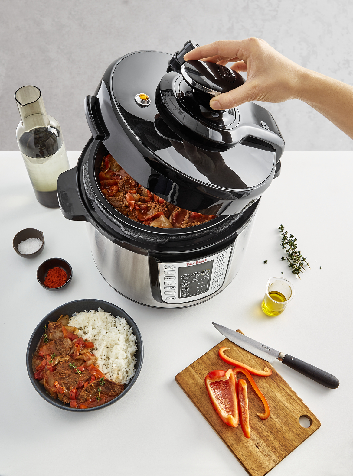 Een persoon opent een zwart-zilveren snelkookpan met rijst en stoofpot, plus ingrediënten.