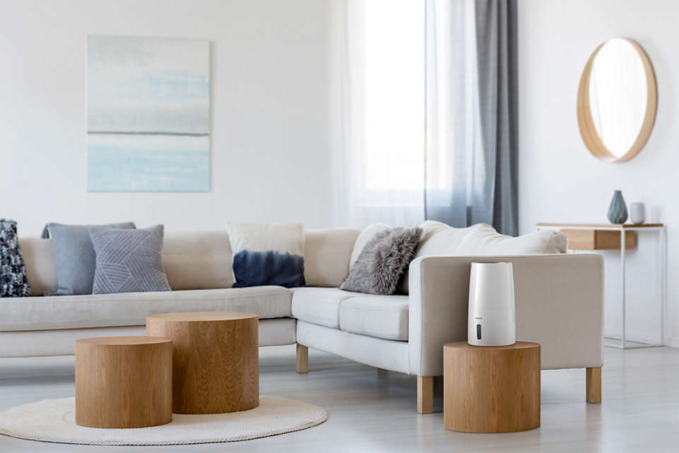 Ein Wohnzimmer mit weißem Sofa, Holztischen und einem Luftbefeuchter rechts. Weiße Wände.