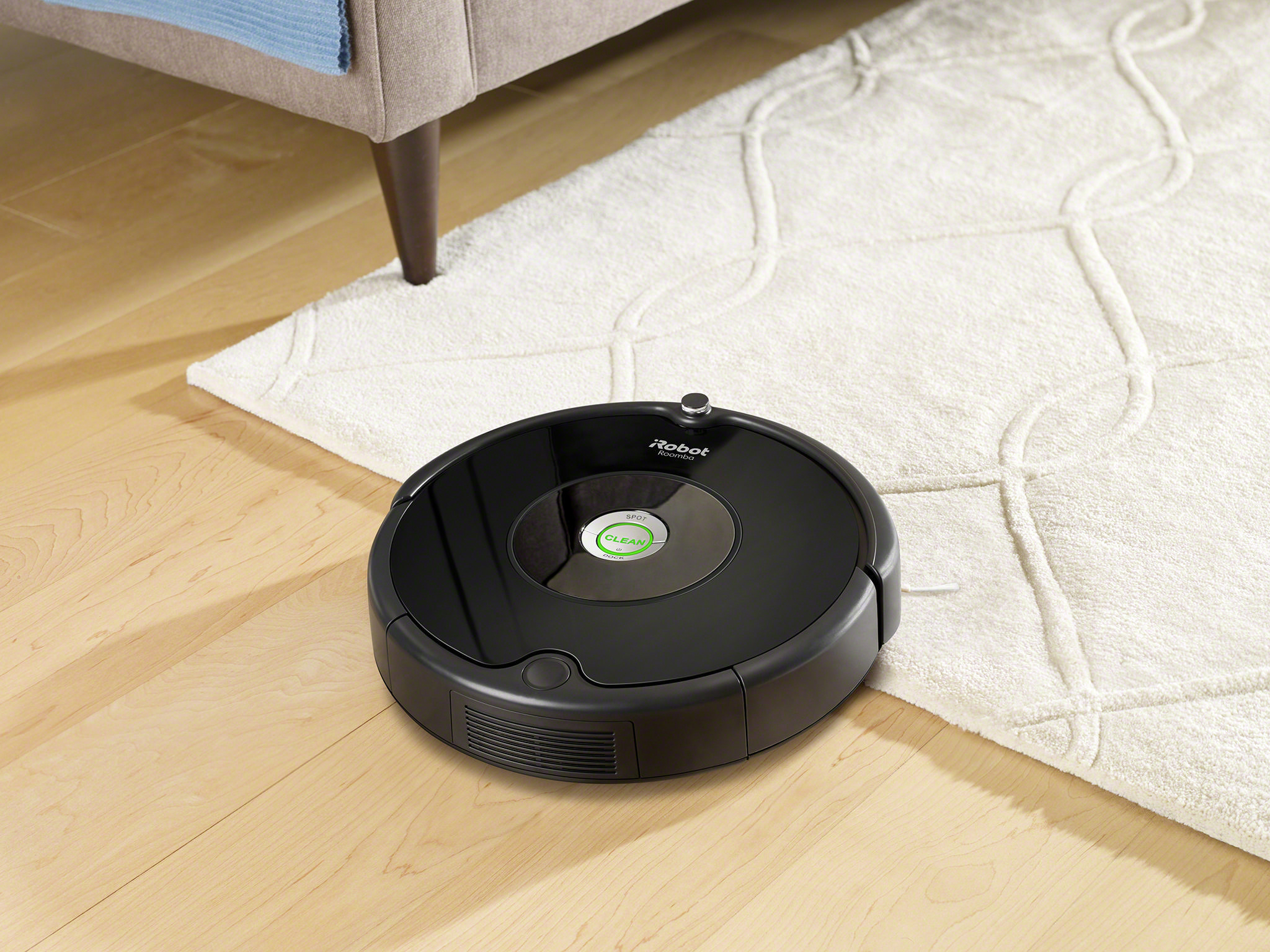 Roboter-Staubsauger auf Holzboden neben einem Teppich. Sofa und Teppich im Bild.