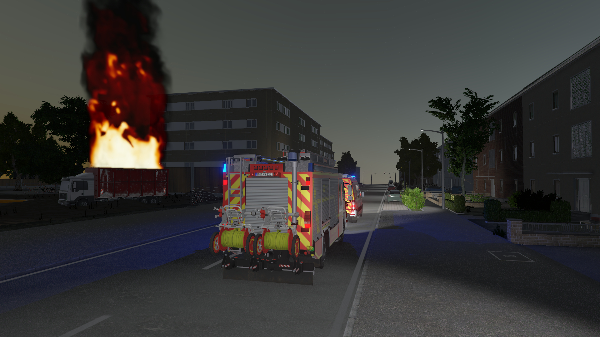 Feuerwehrfahrzeuge auf einer Straße bei Nacht mit brennendem Gebäude im Hintergrund.