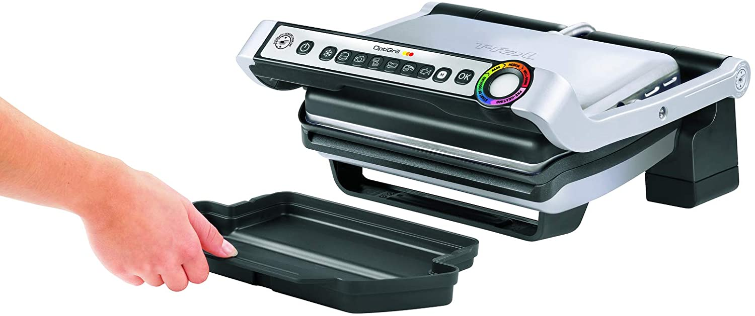 Una mano tiene un vassoio nero vicino a un T-Fal OptiGrill. La griglia è argentata e nera con pannello di controllo e indicatore di cottura.