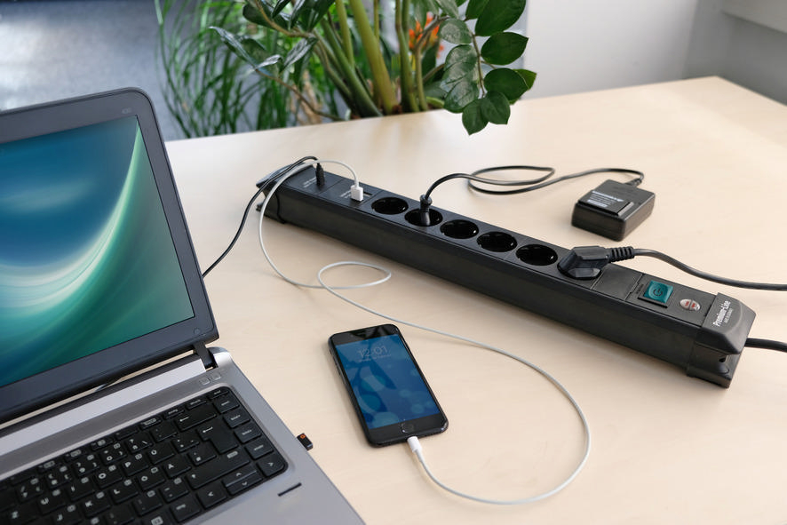 Ein Laptop, ein ladendes Telefon und eine Steckdosenleiste auf einem hellen Holztisch.
