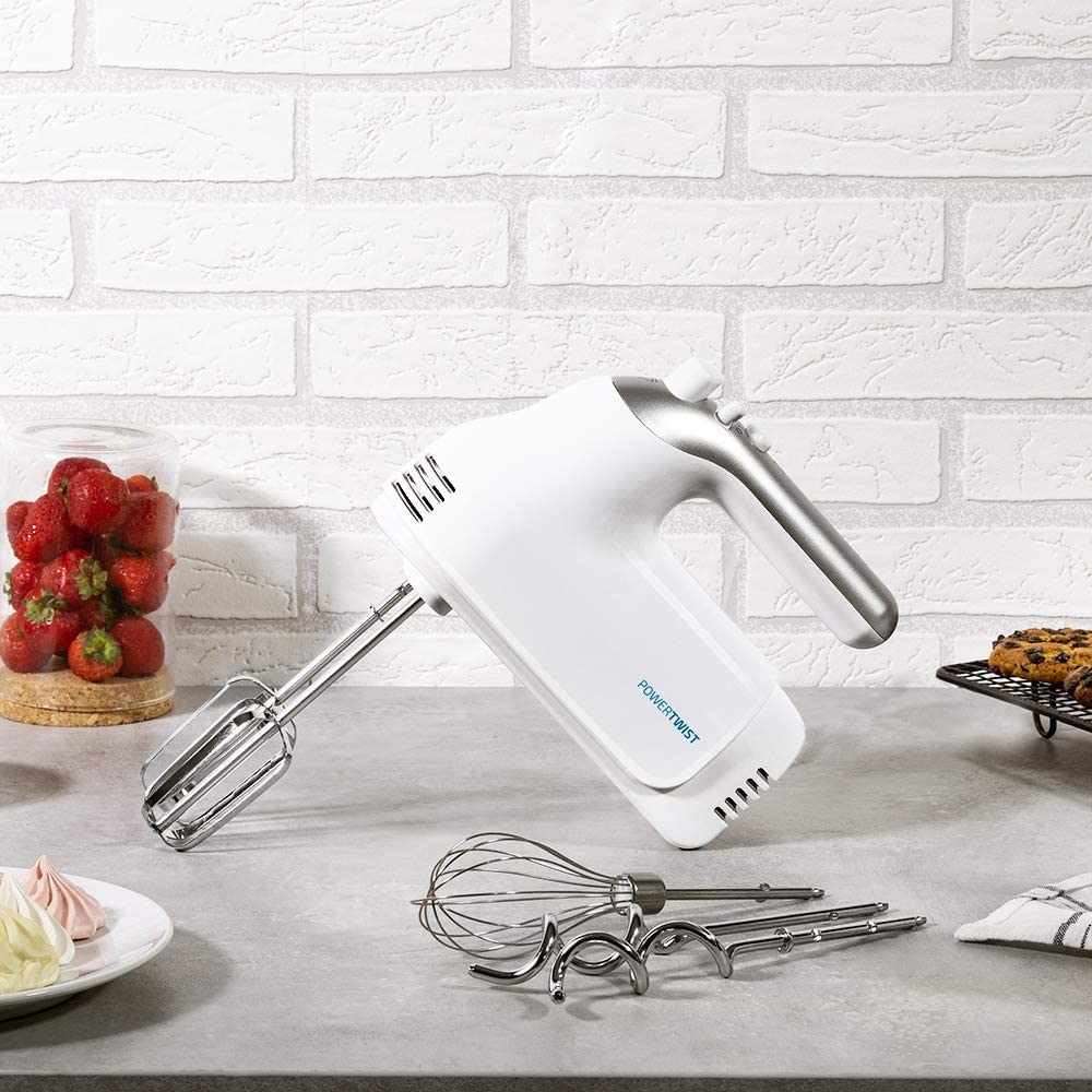 Batidora de mano blanca con accesorios y fresas. Pared de ladrillo al fondo.