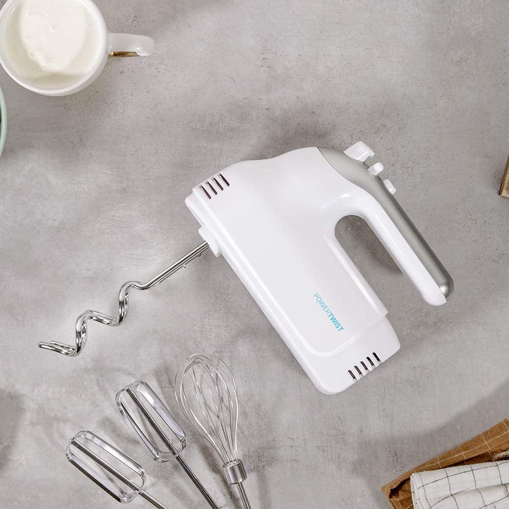 Batidora de mano blanca con accesorios sobre una superficie gris, con una taza de leche.