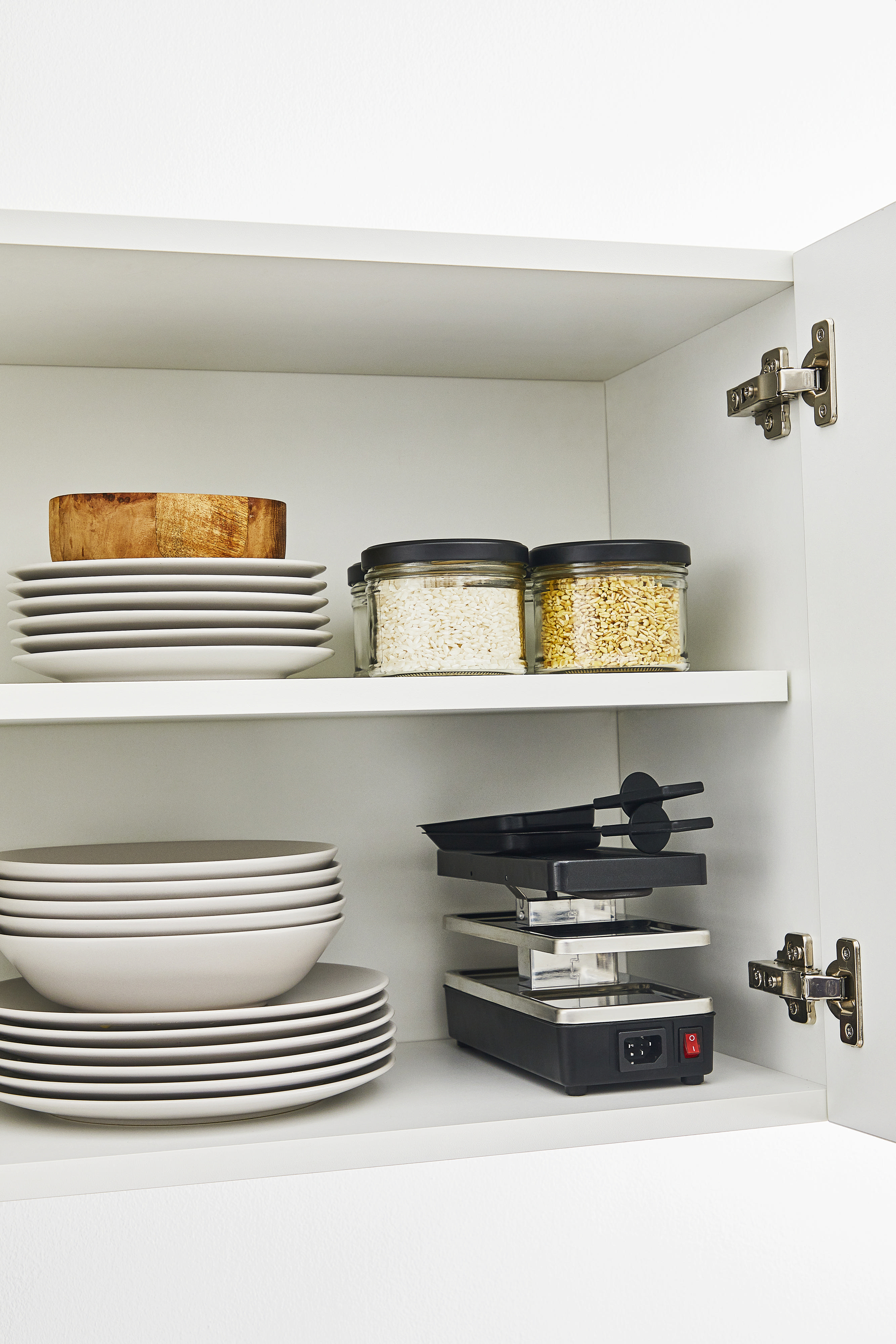 Ein weißer Schrank mit offenen Türen, Regalen mit Tellern, Gläsern und einem Raclette-Grill. Gut organisierte Küche.