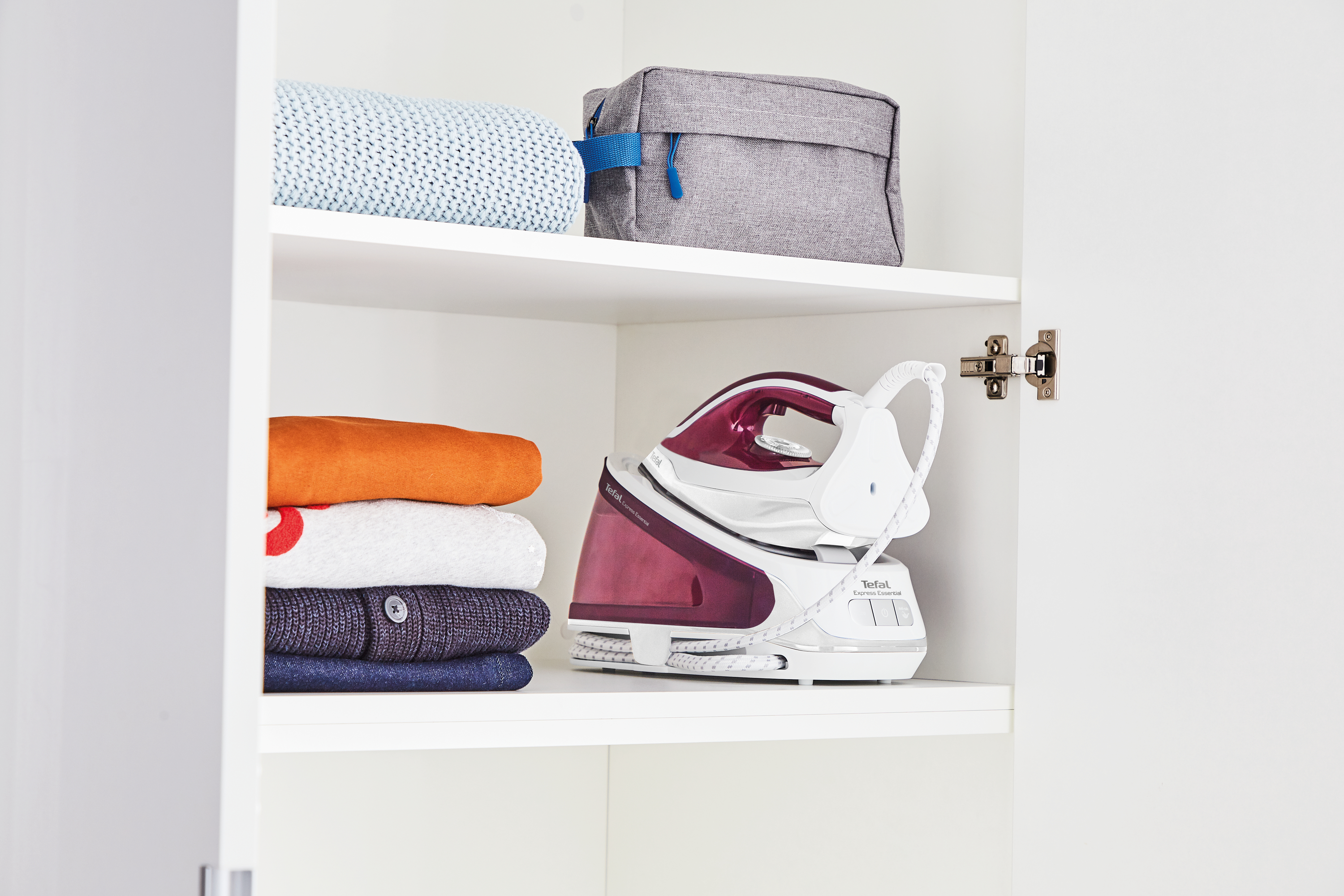 Weißer Schrank mit Kleidung, Bügeleisen und grauer Kosmetiktasche. Das Bügeleisen ist rot-weiß.