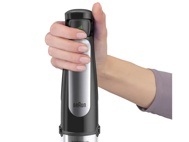 Egy kéz egy fekete-ezüst Braun botmixert tart. A mixer zöld fényekkel van bekapcsolva.