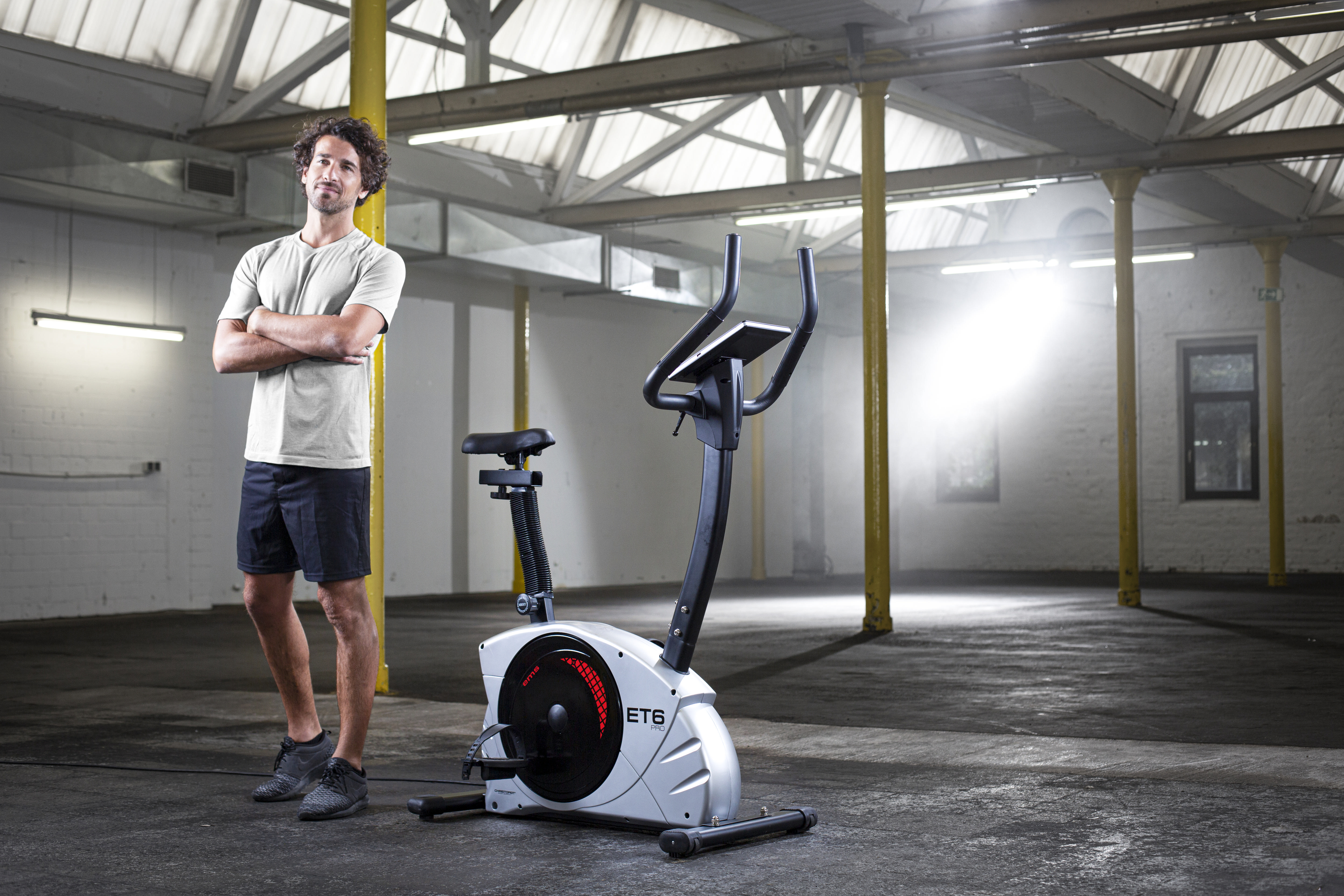 Mann mit verschränkten Armen steht neben einem Heimtrainer in einem großen, leeren Lagerraum.