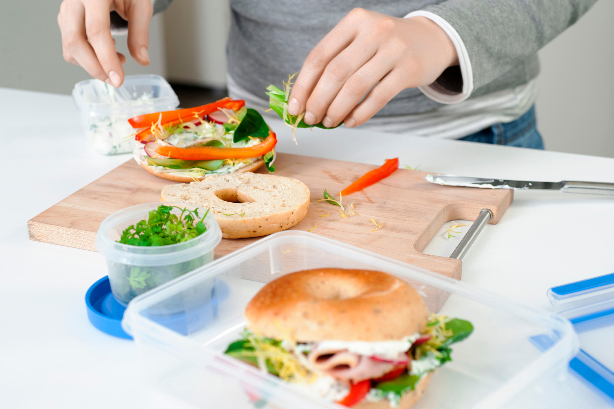 Person bereitet ein Bagel-Sandwich mit Salat, Tomaten und anderen Zutaten auf einem Holzbrett zu.