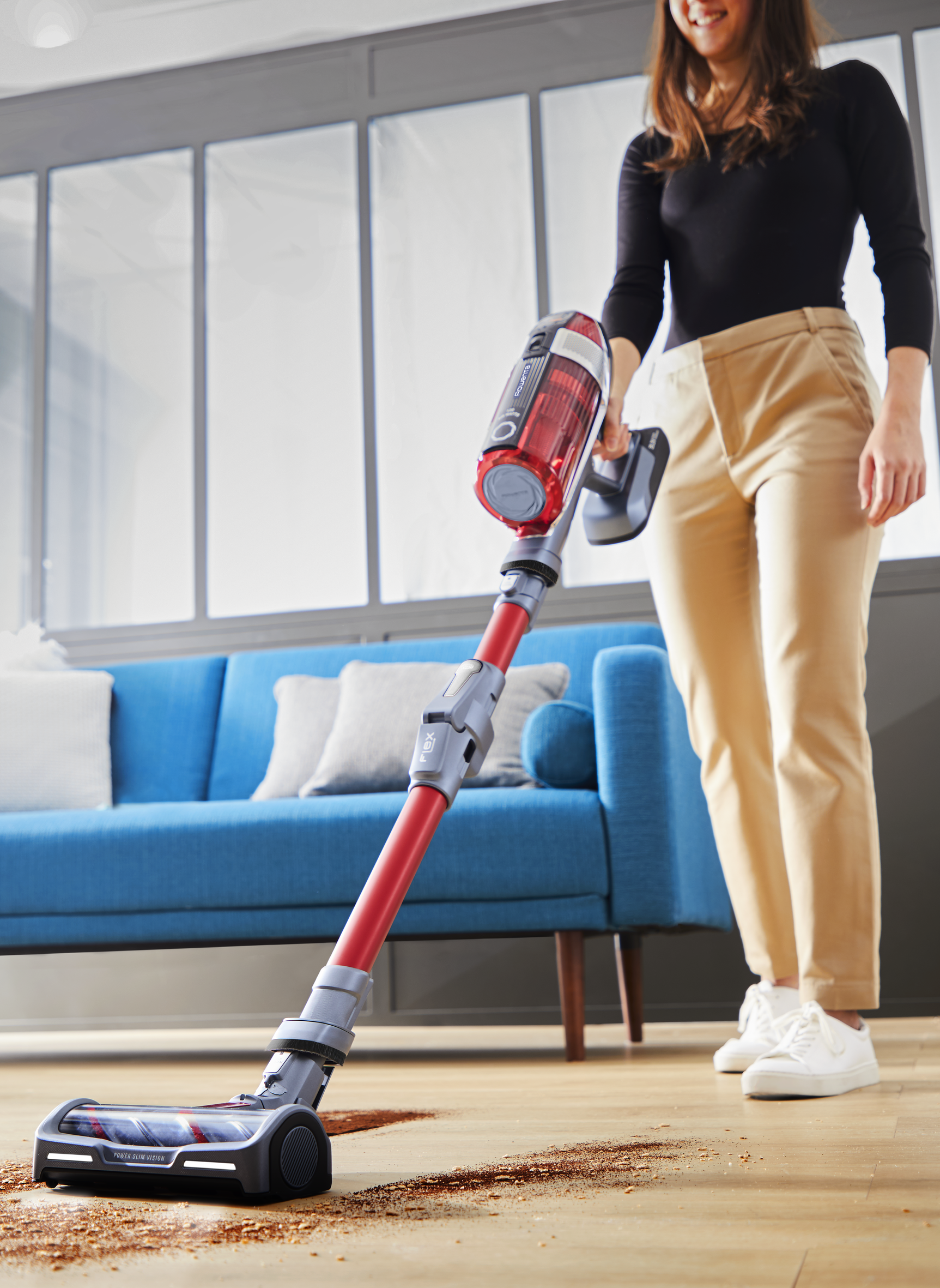 Une femme passe l'aspirateur dans un salon avec un aspirateur sans fil rouge et gris.