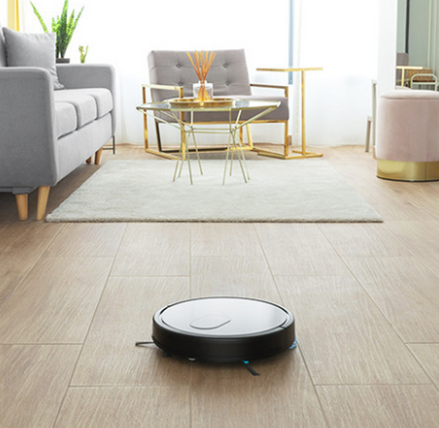 Una aspiradora robot negra y plateada en un suelo de madera. Hay una alfombra y muebles.