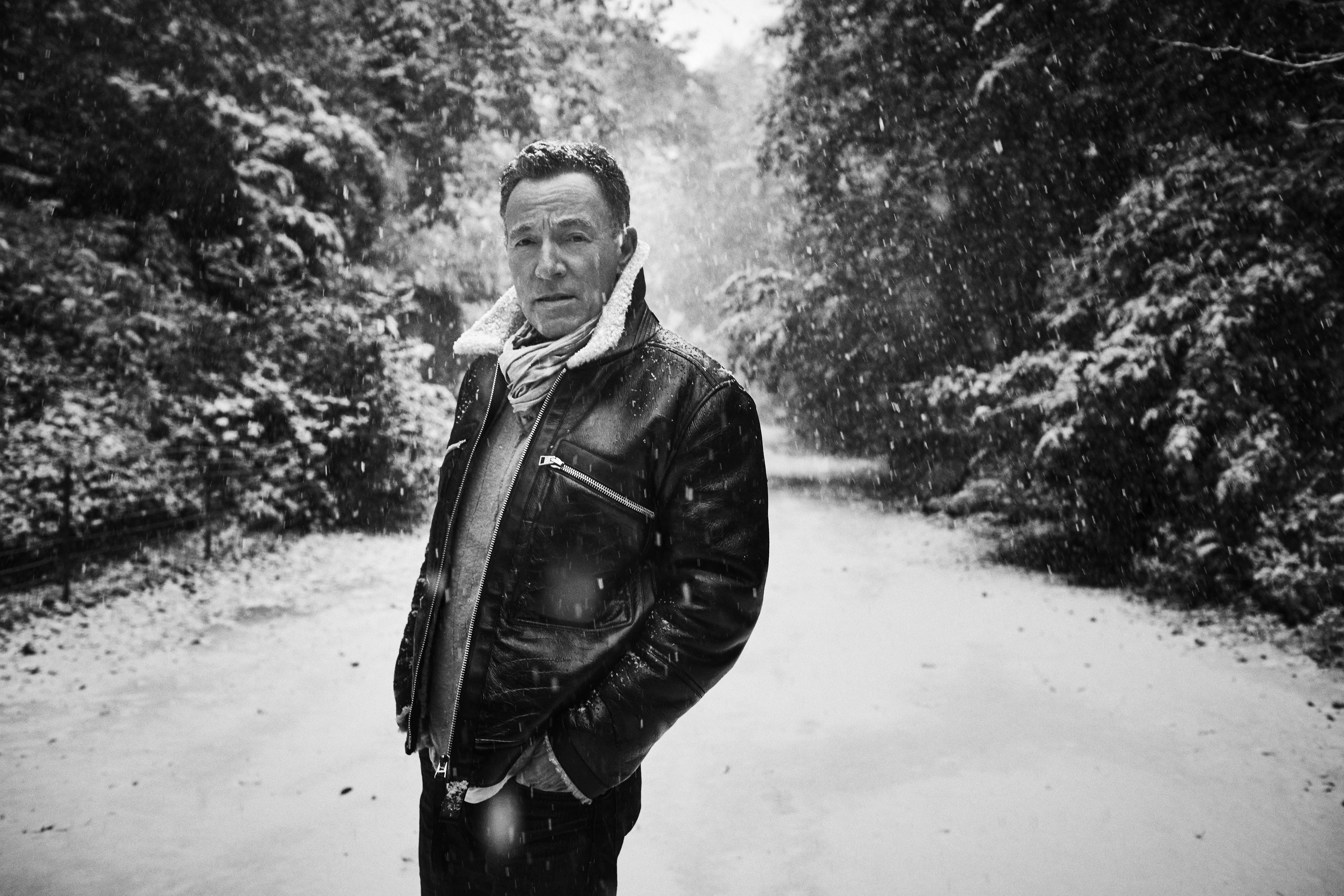 Mann in schwarzer Jacke steht auf verschneitem Weg, Bäume an Seiten, Schnee fällt.
