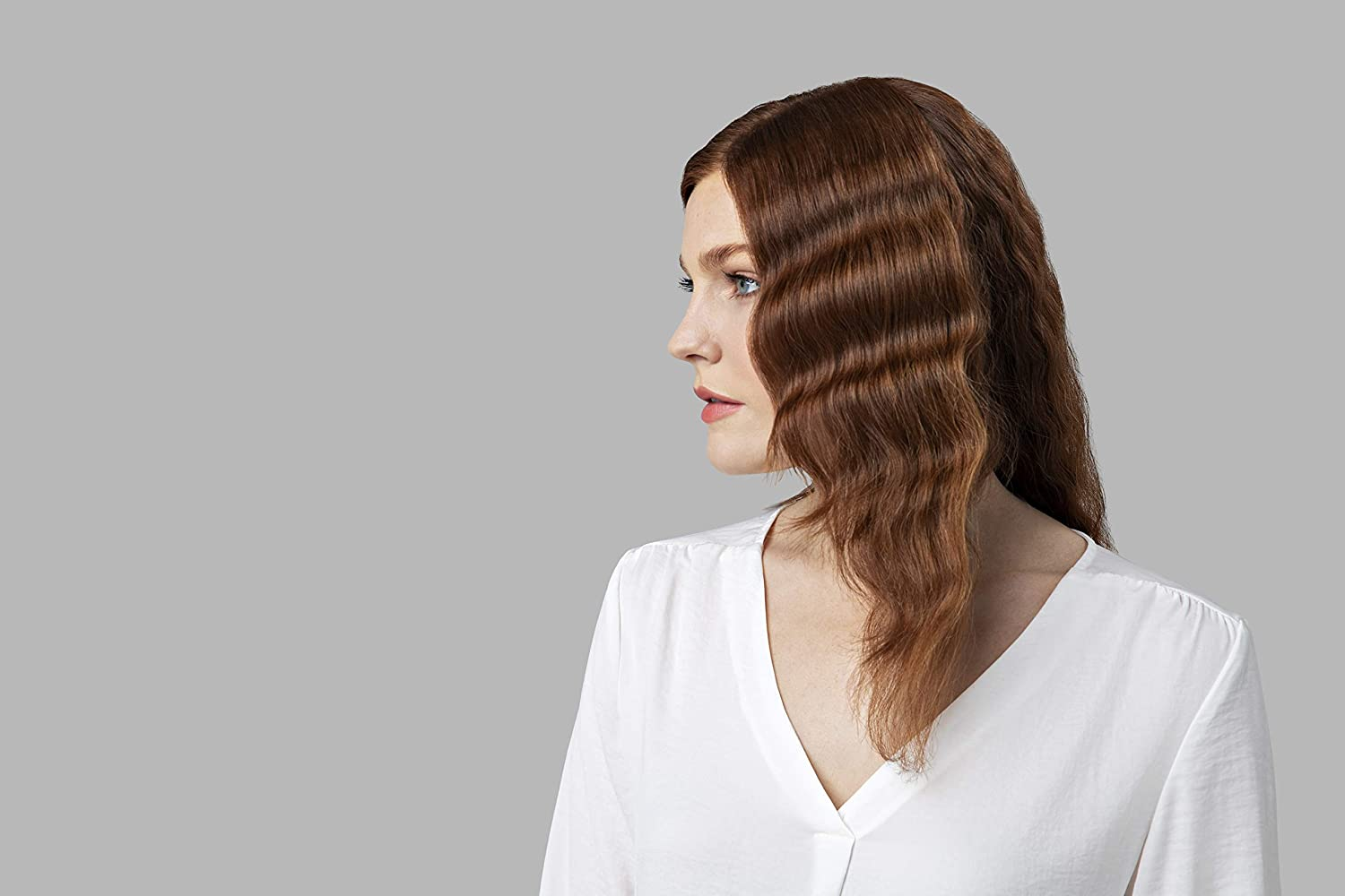 Eine Frau mit roten welligen Haaren und einem weißen Hemd blickt auf hellem grauem Hintergrund nach links.