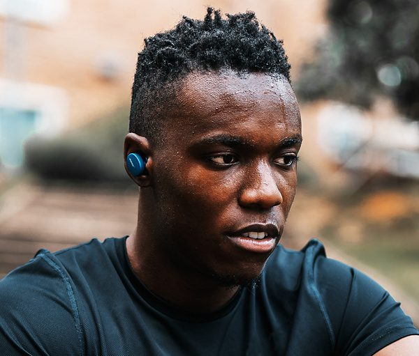 Un homme noir en sueur, aux cheveux courts, foncés et frisés, porte un écouteur bleu et une chemise sombre.