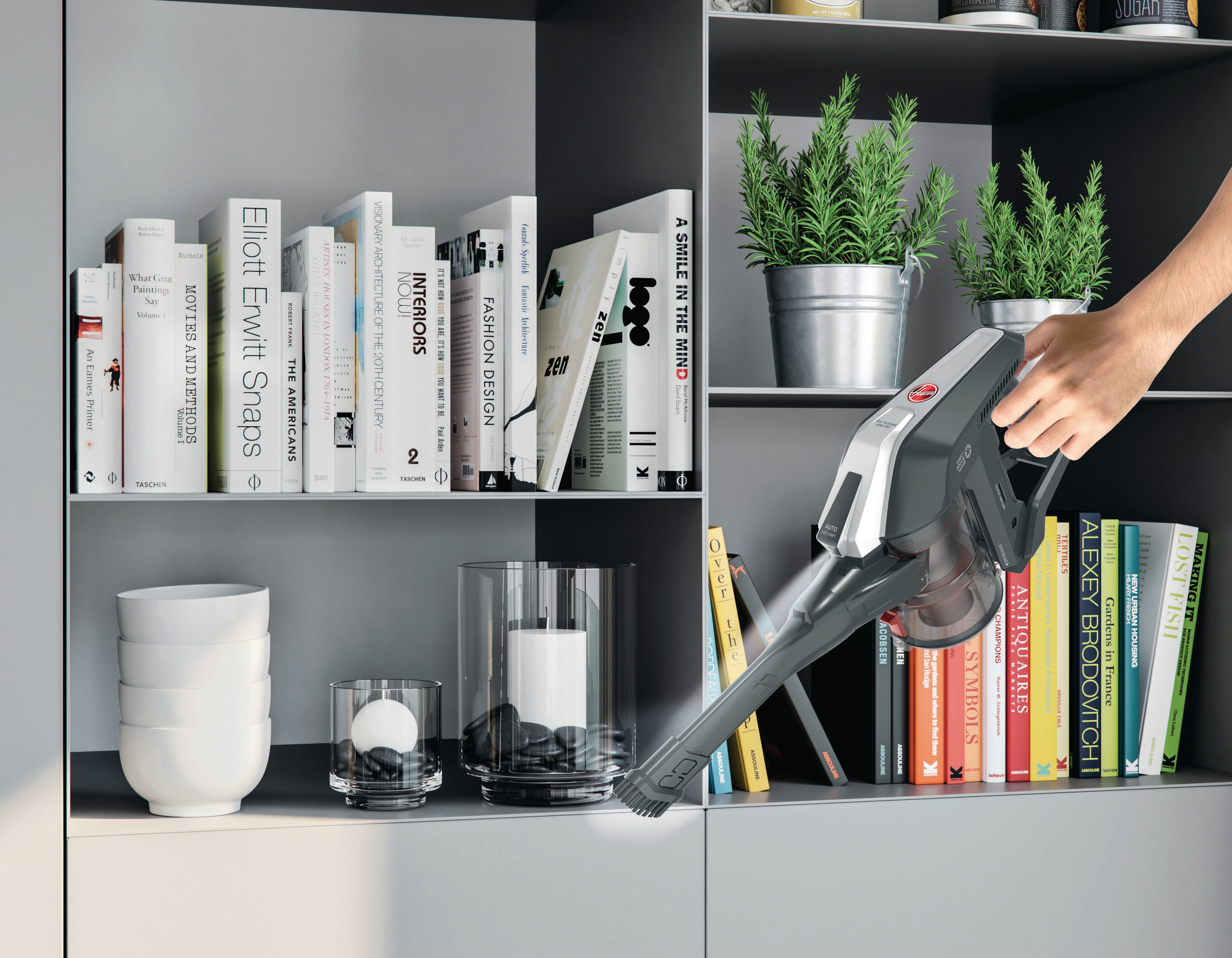 Hand hält grauen und roten Staubsauger und reinigt ein weißes Regal mit Büchern und Pflanzen.