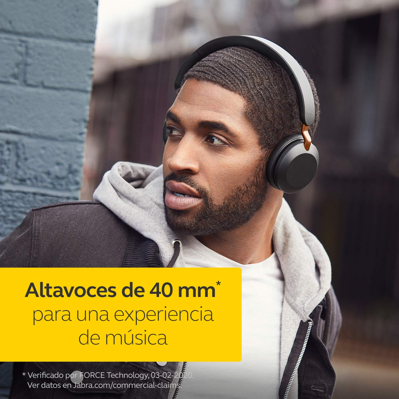 Hombre con auriculares negros, sudadera gris, mirando de lado contra pared ladrillos.