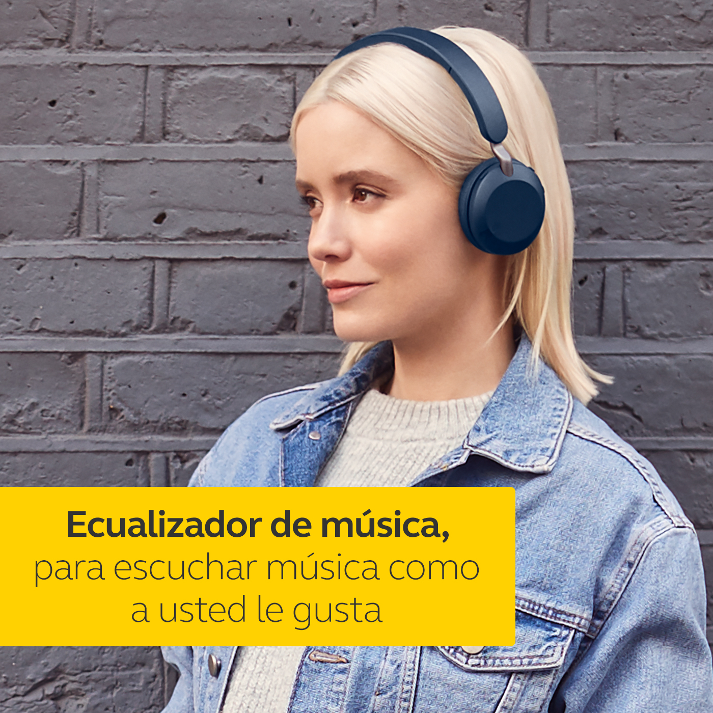 Mujer con auriculares azules, chaqueta vaquera, mirando hacia otro lado contra pared de ladrillos.