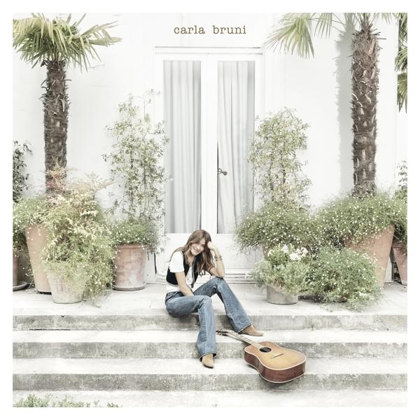 Carla Bruni zit op treden met gitaar. Palmbomen en planten op de achtergrond. Wit huis.