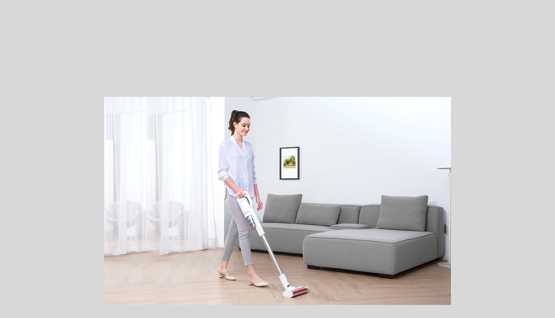 Eine Frau saugt einen Raum mit einem weißen kabellosen Staubsauger. Ein graues Sofa und Bild sind sichtbar.