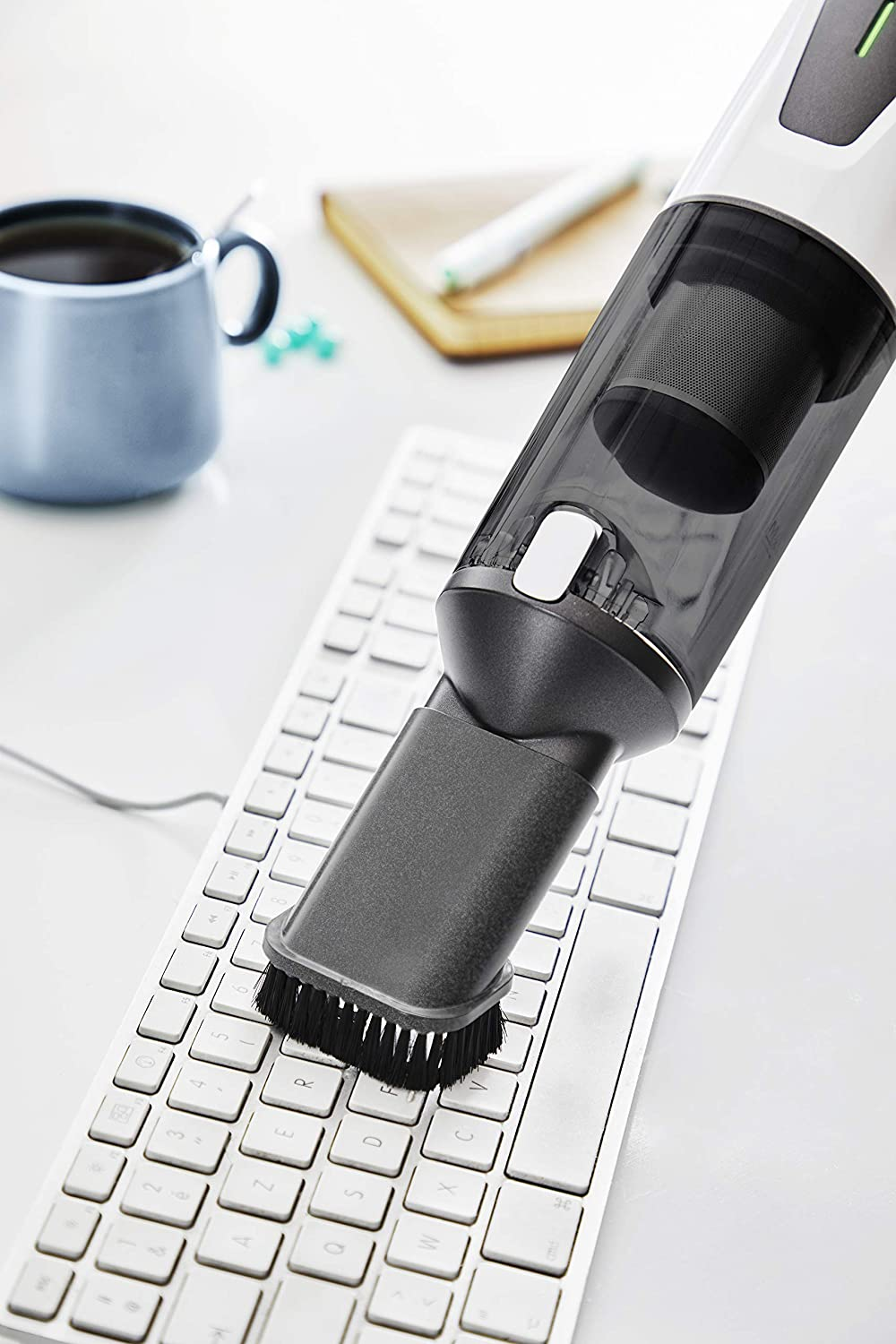 Ein Handstaubsauger reinigt eine Computertastatur. Ein weißer Schreibtisch und eine blaue Tasse sind im Hintergrund.