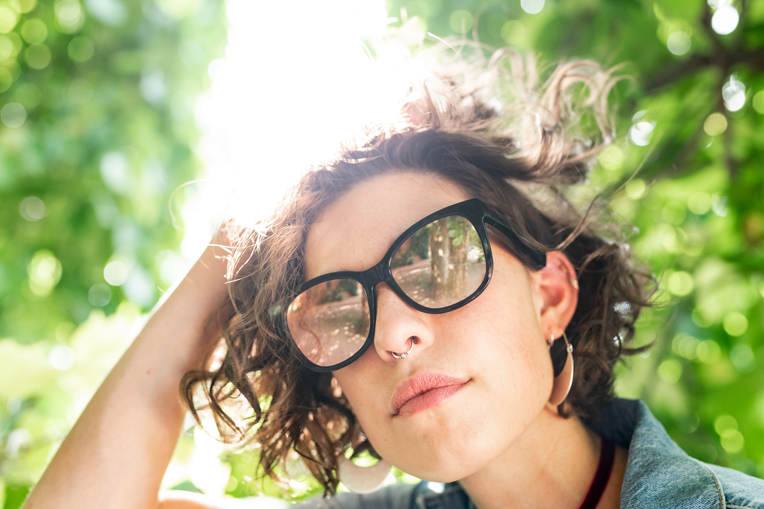 A woman wearing sunglasses with reflection, poses under the sunlight with green trees.