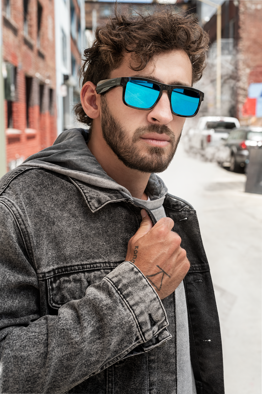 Man wearing sunglasses with blue lenses, denim jacket, and a tattoo, outdoors.