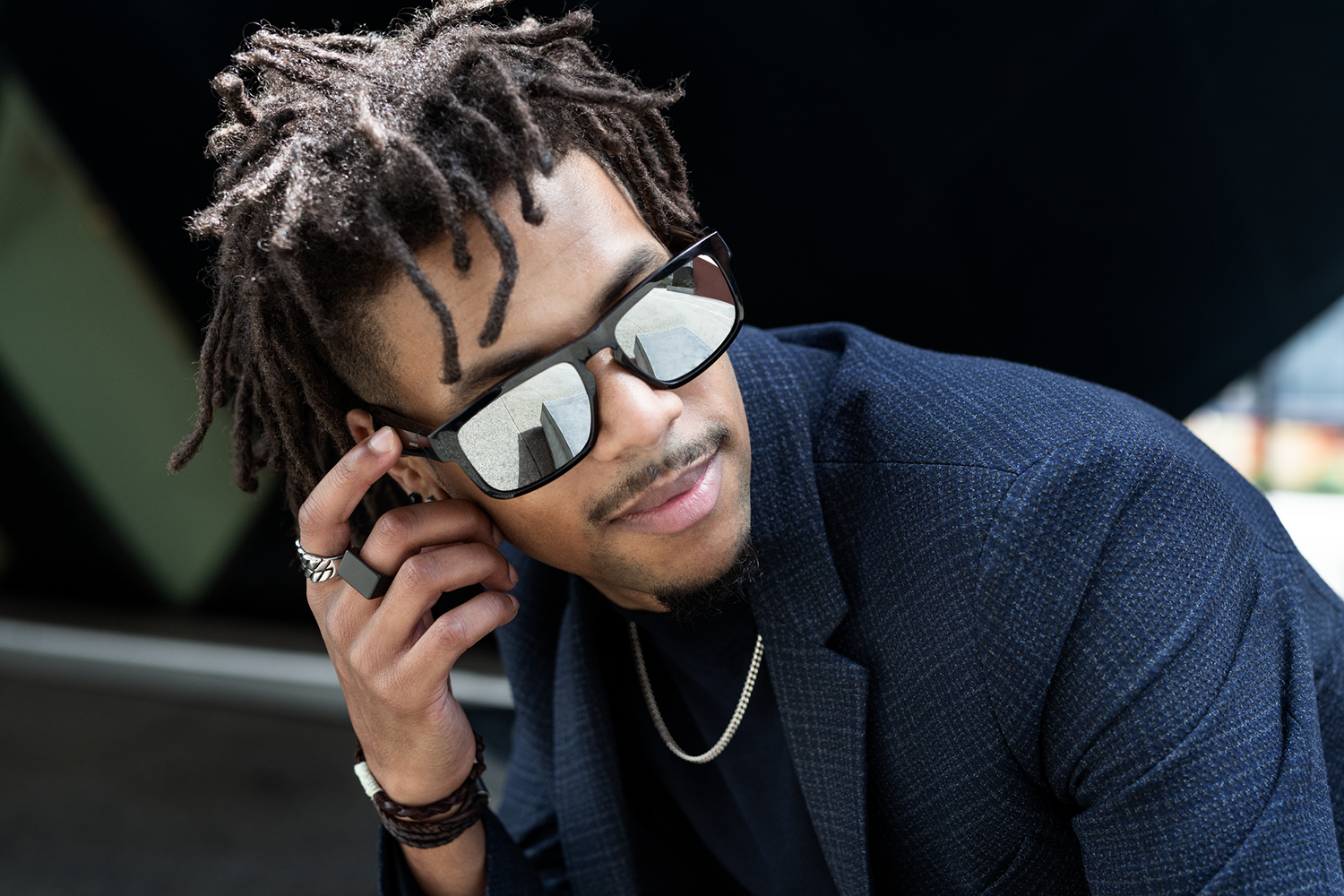 A man with dreadlocks wears sunglasses and a blue blazer. He is touching the glasses with his finger. A silver chain is visible.