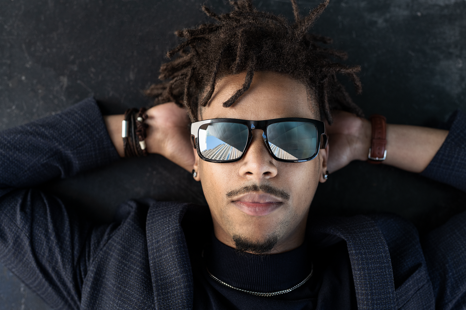 A man with dreadlocks wearing sunglasses and a suit, resting his hands behind his head. The sunglasses reflect a building.