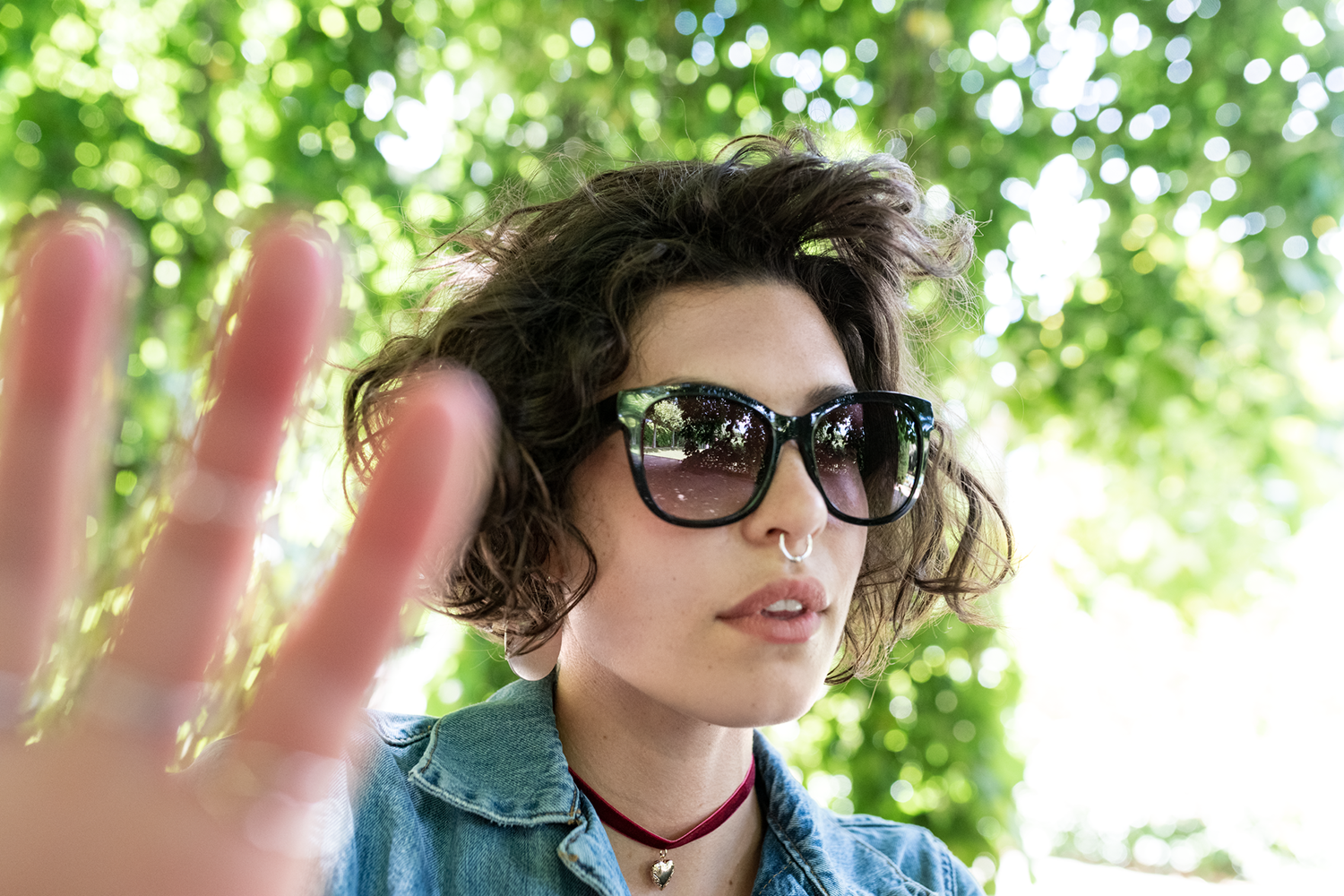A woman wearing sunglasses and a denim jacket extends her hand towards the camera. Green foliage forms the background.