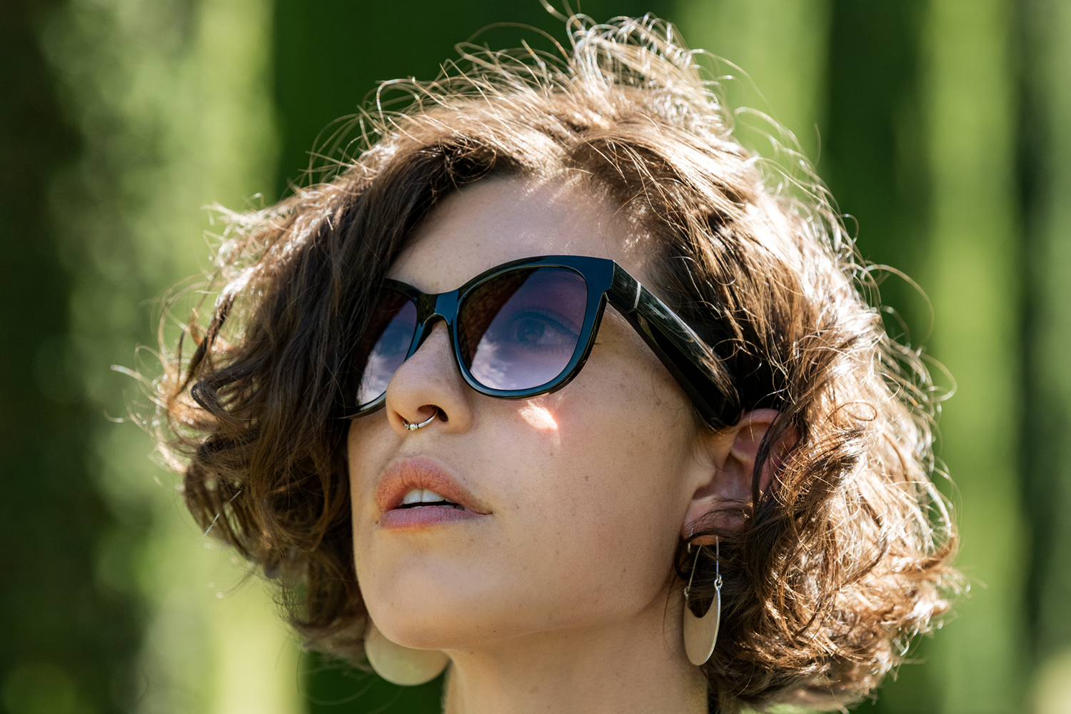 A woman with sunglasses and curly brown hair. She has a nose ring and earrings. Green background.