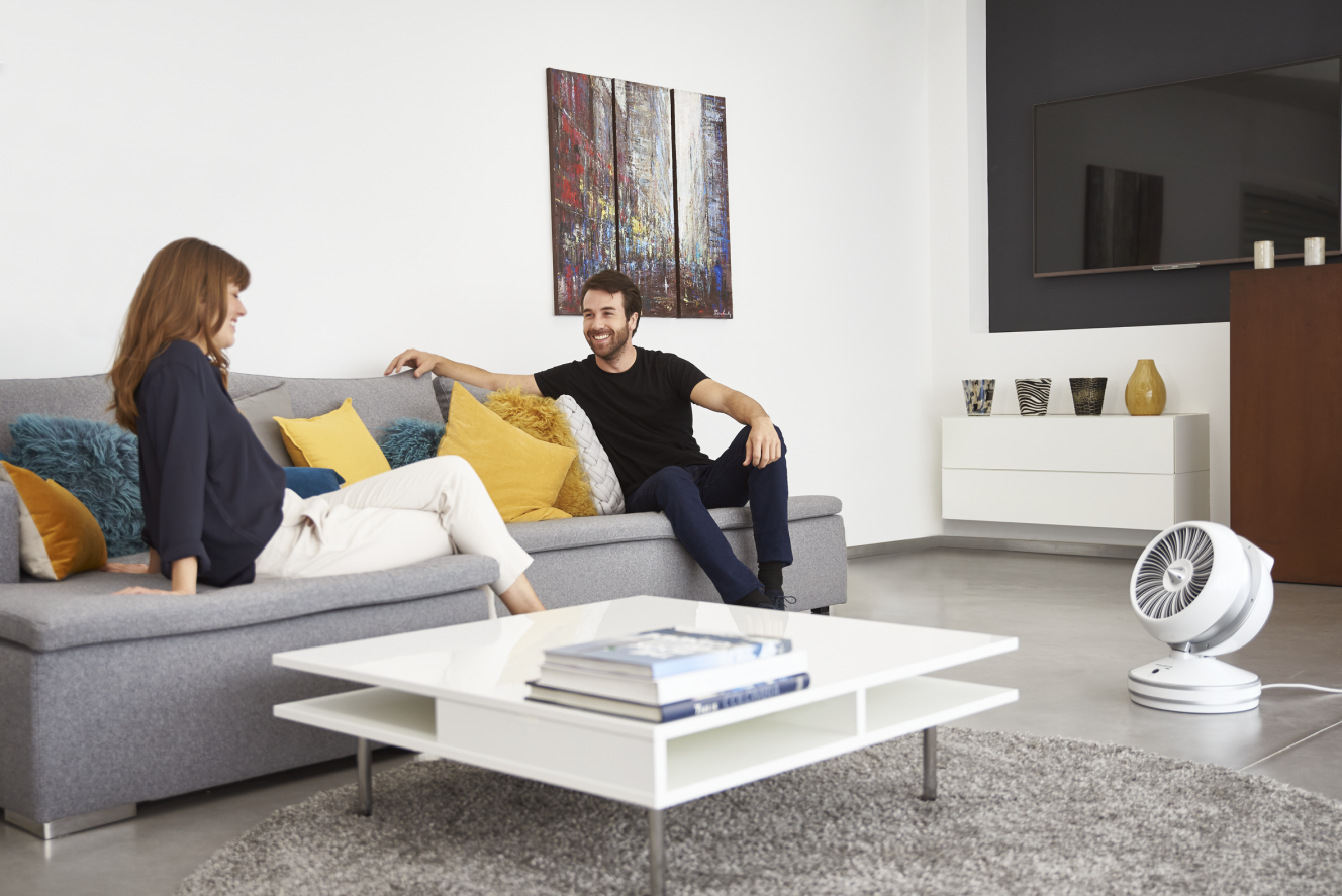 Couple sur un canapé gris, table blanche avec des livres et un ventilateur blanc dans le salon.