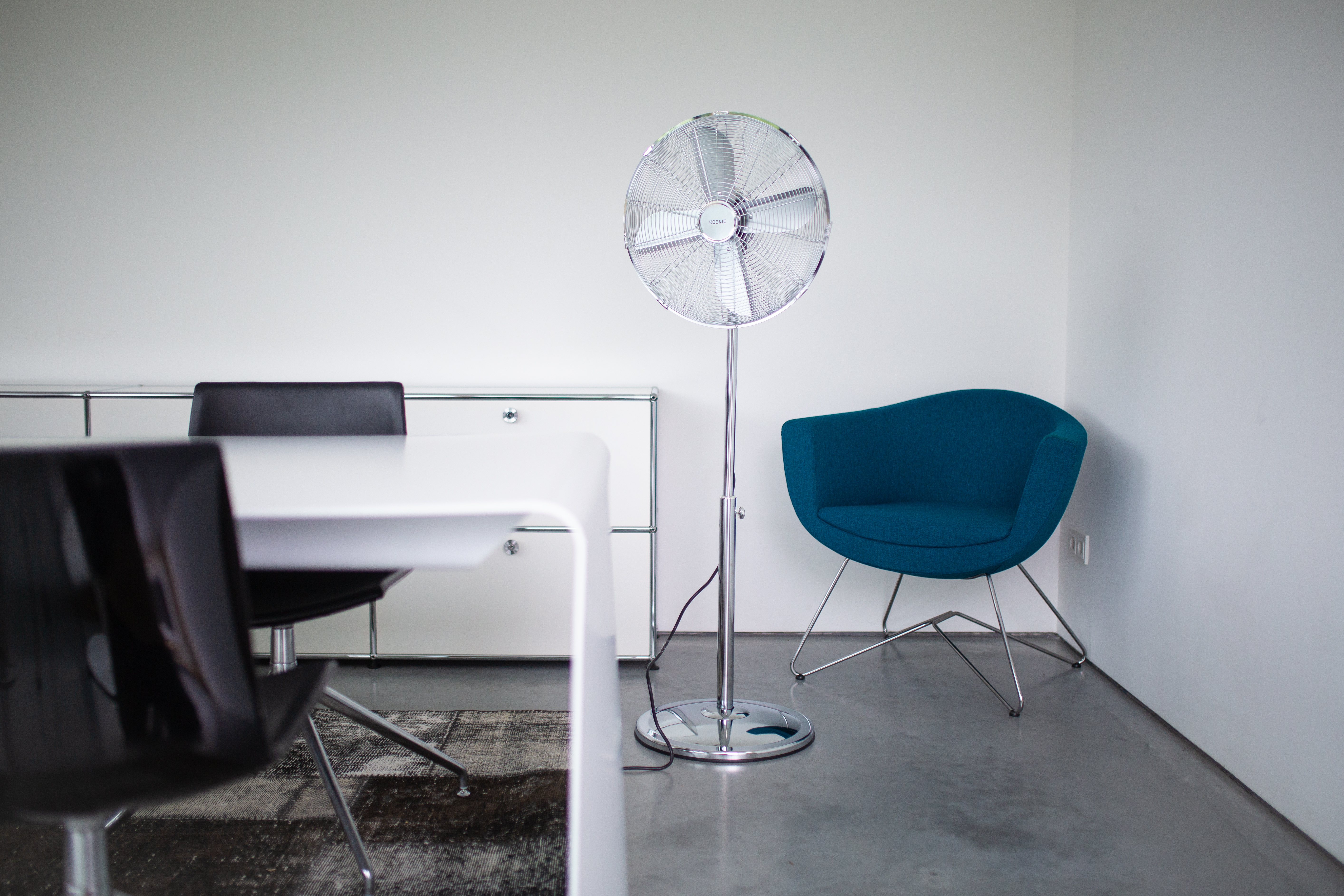Un ventilatore rotondo si trova accanto a una sedia blu e un tavolo bianco.