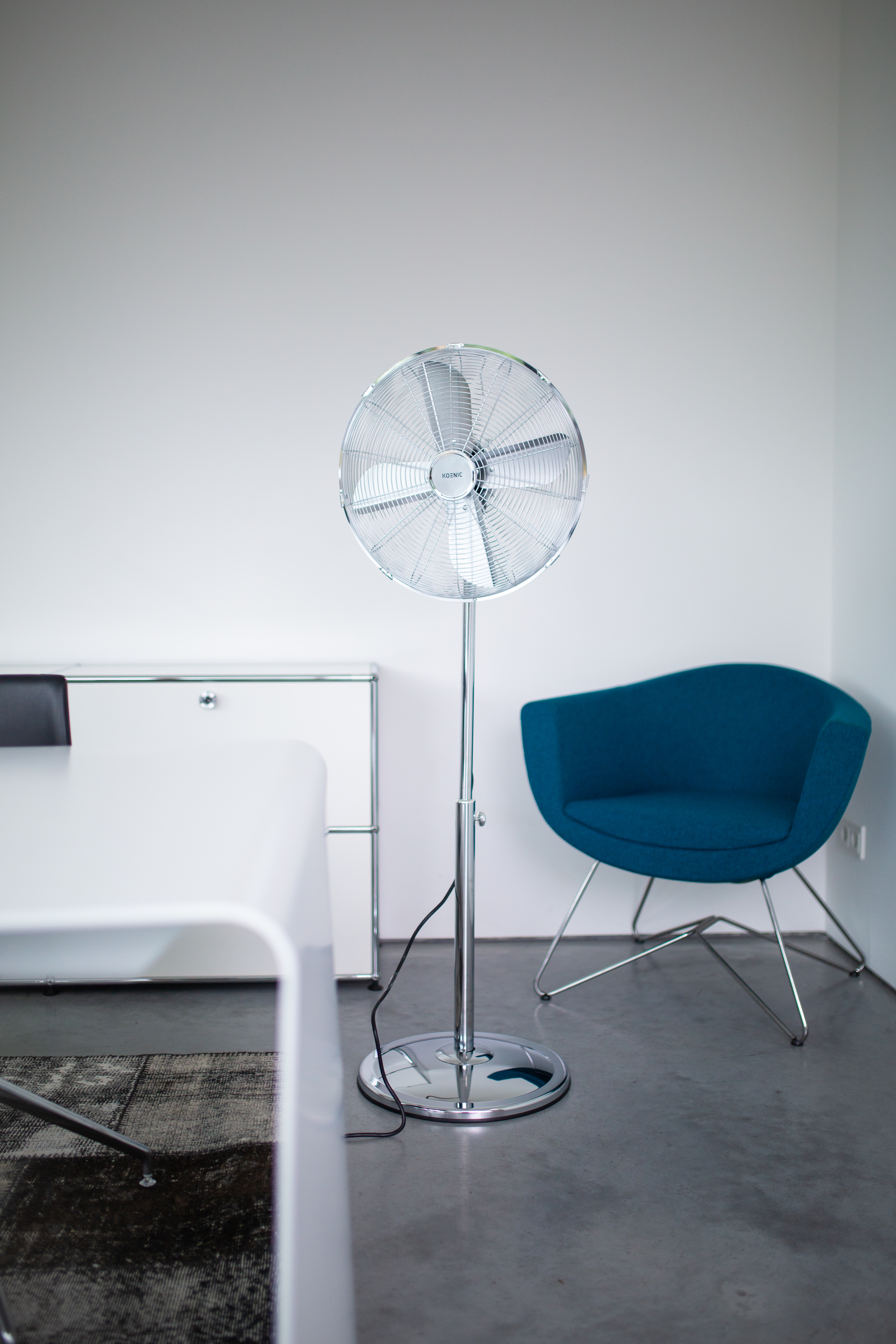 Un ventilatore da terra argentato è accanto a una poltrona blu e un mobile bianco.