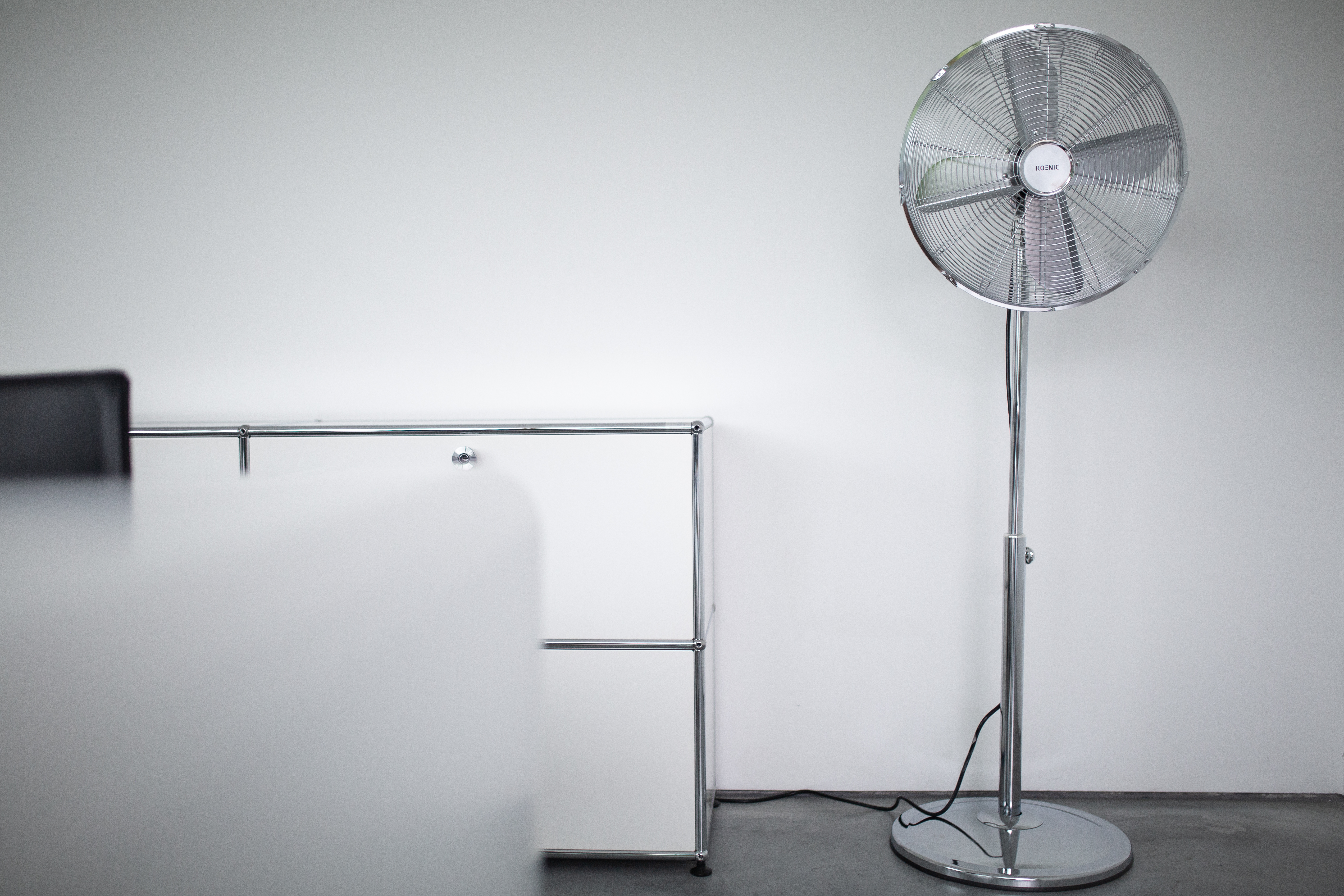 Un ventilatore da terra argentato si trova accanto a un mobile bianco contro un muro bianco.