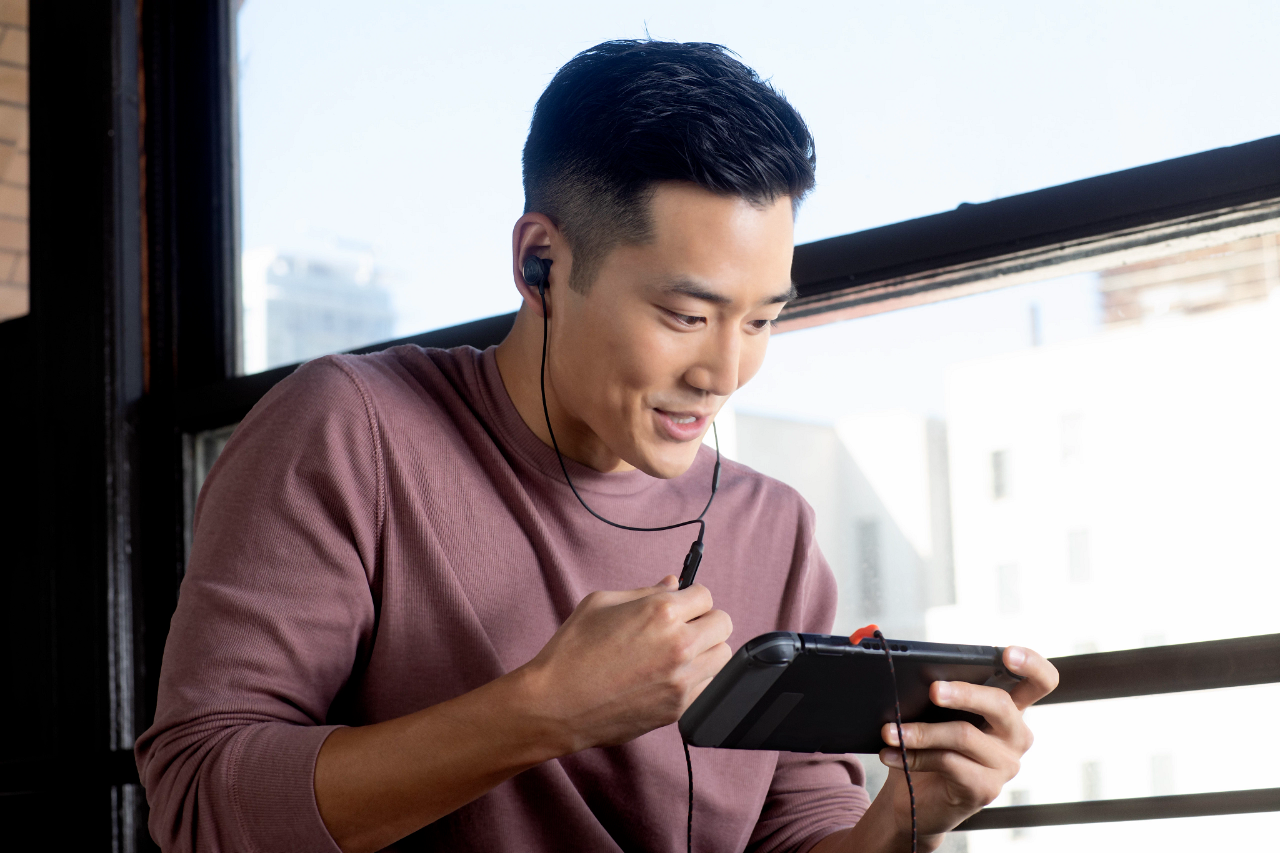 A man is playing a video game on a handheld device with earbuds. He is smiling.