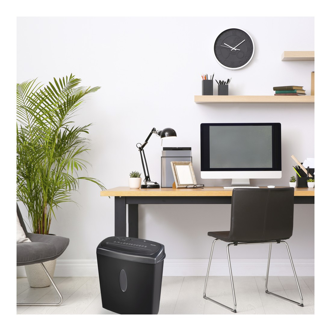 Büro mit Schreibtisch, Computer, Pflanze und einem schwarzen Aktenvernichter. Stuhl und Regale sind ebenfalls sichtbar.