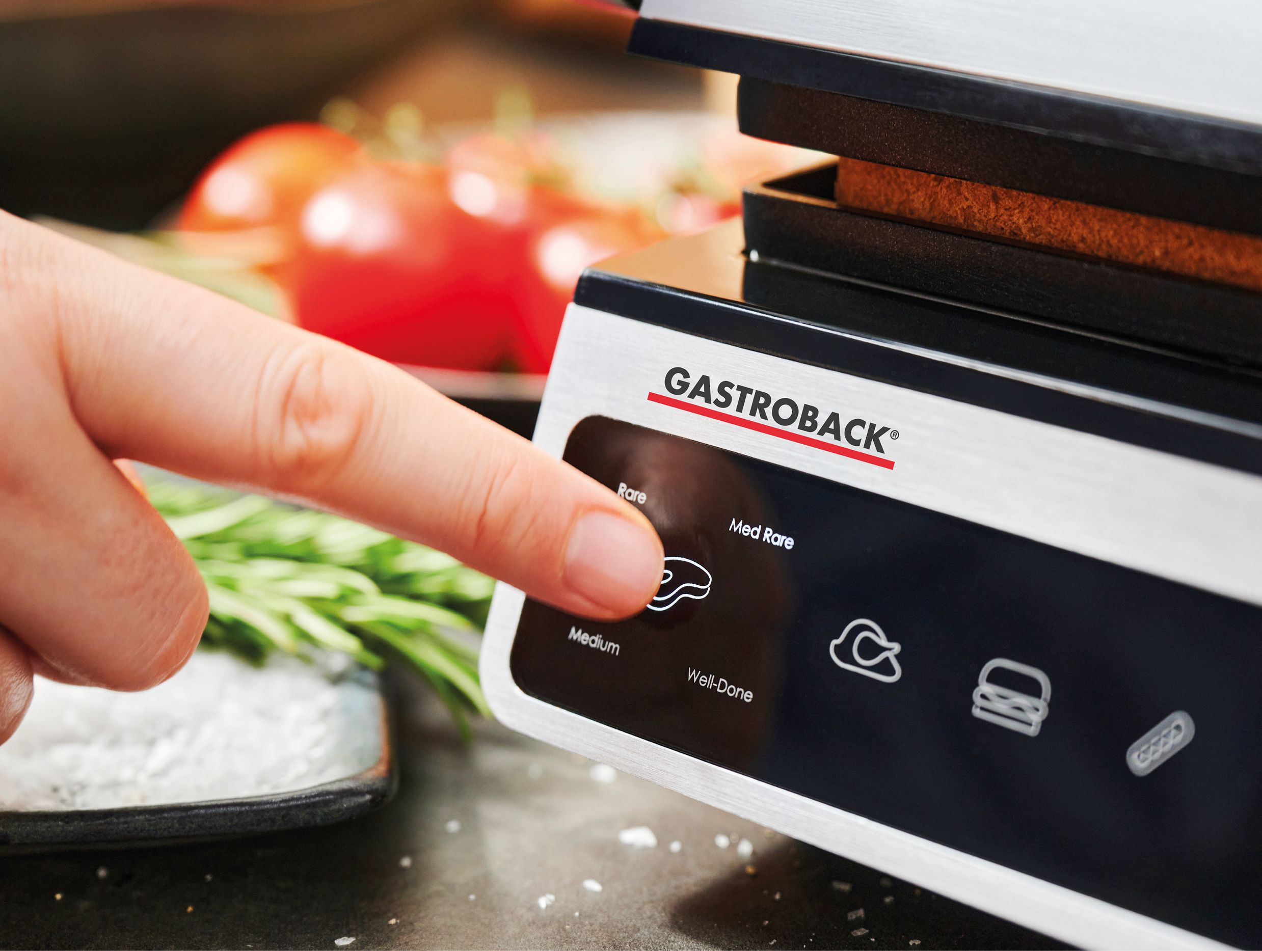 Ein Finger drückt eine Taste auf einem Gastroback-Grill, mit Tomaten und Rosmarin im Hintergrund.