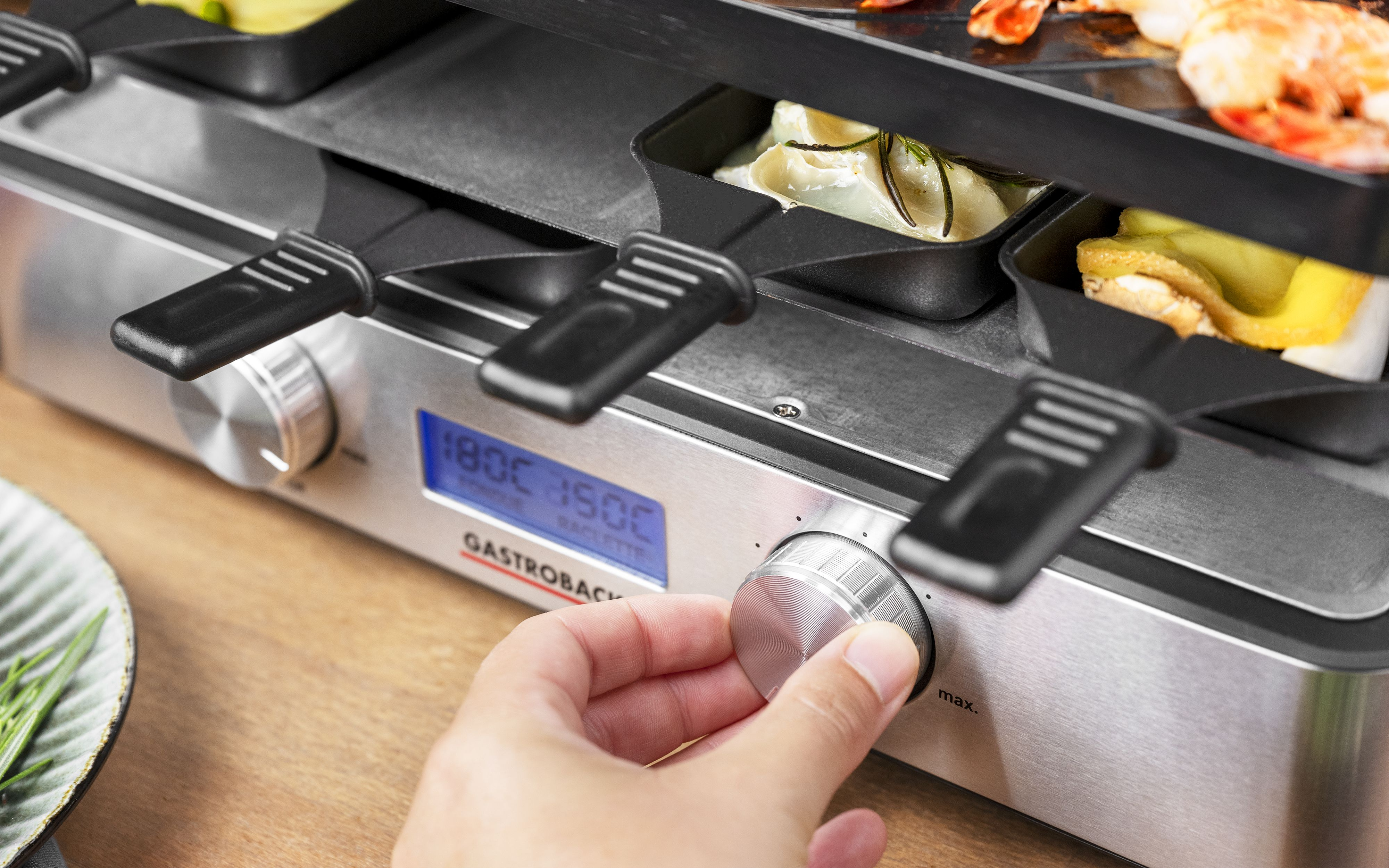 Die Hand einer Person, die einen Drehregler an einem silbernen Raclette-Grill mit Essen einstellt.