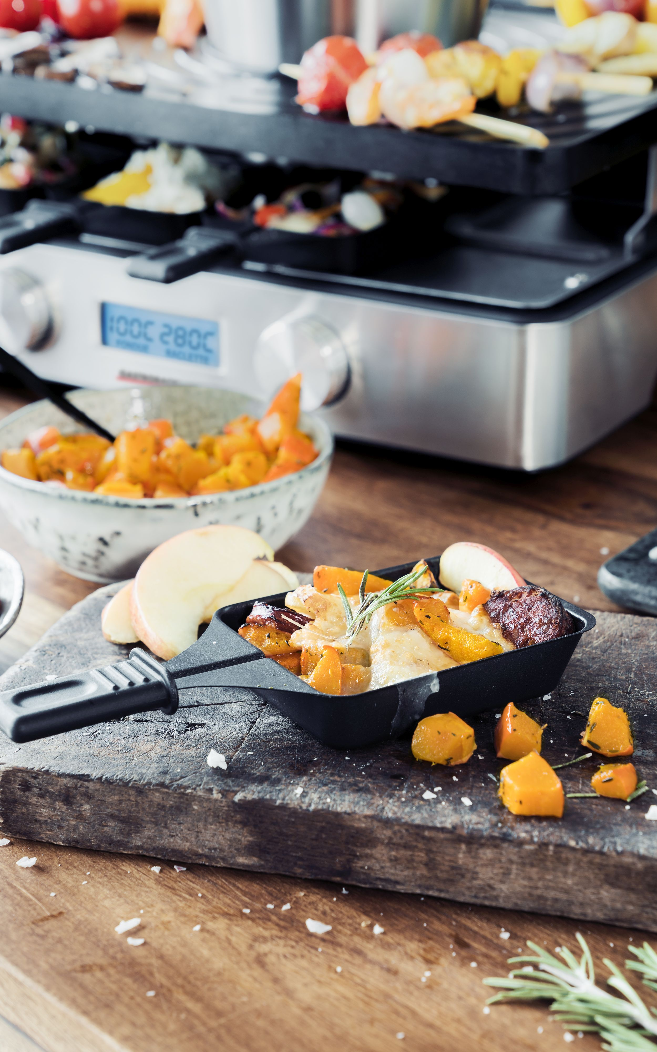 Raclette-Gericht mit gegrilltem Essen auf einem Holzbrett, mit Beilagen und Grill im Hintergrund.