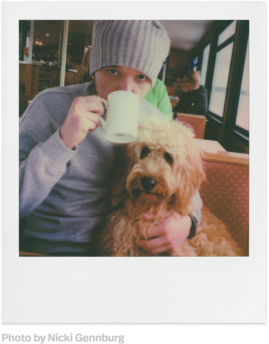 Eine Person hält eine weiße Tasse, während sie neben einem Hund auf einem Sofa sitzt. Es ist ein Polaroid-Foto.
