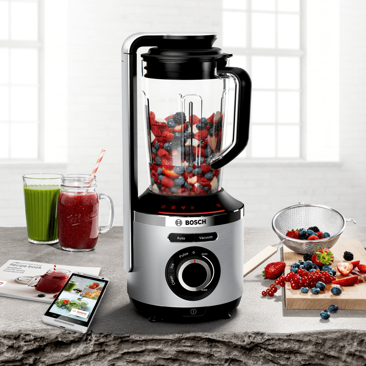 Ein Bosch-Mixer mit Früchten darin. Daneben ein Smoothie und ein Rezeptbuch mit einem Telefon. Beeren, ein Sieb und ein Holzbrett.